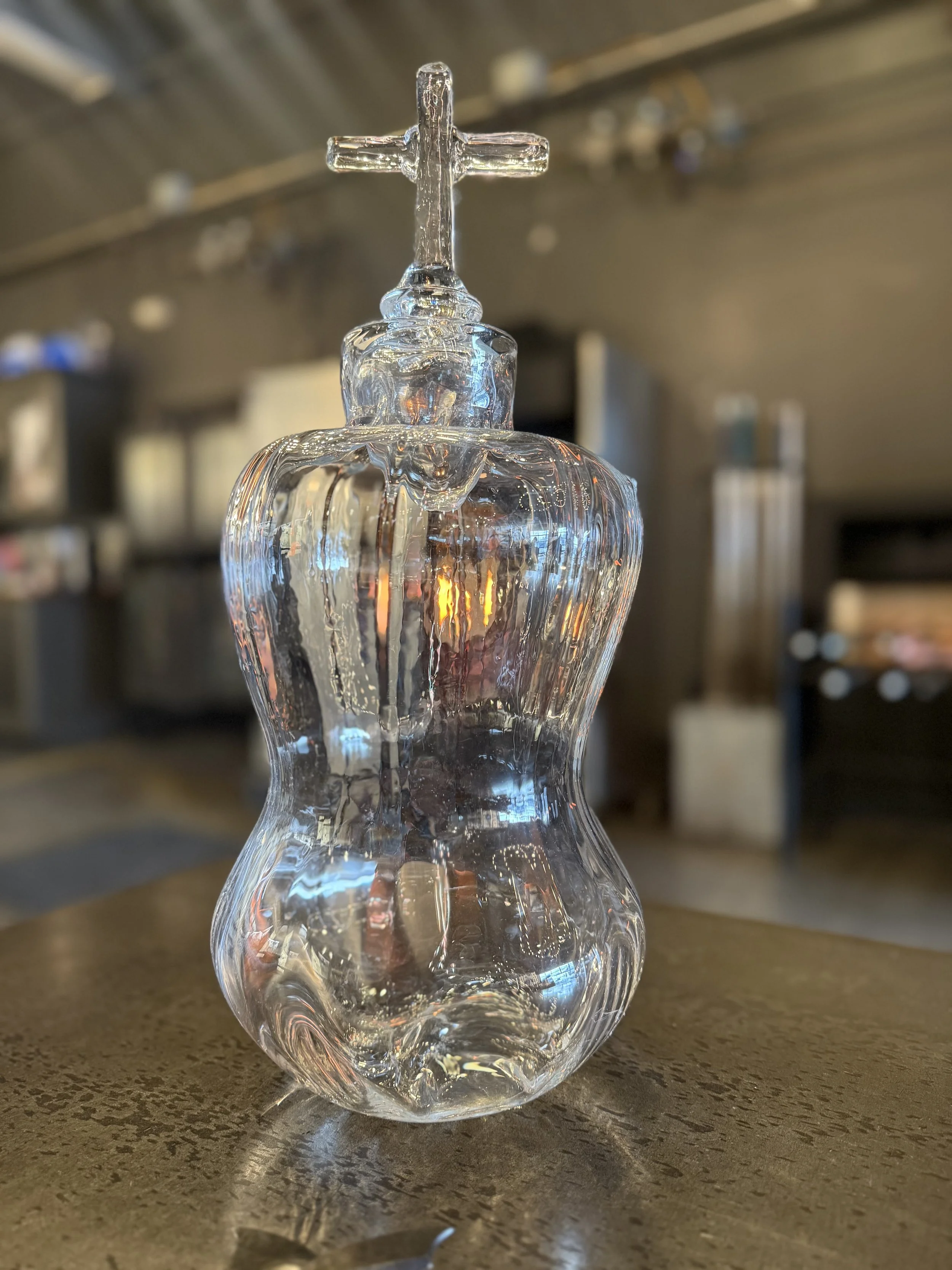 A clear glass decanter with a cross on top, placed on a wooden surface in a blurry indoor setting.