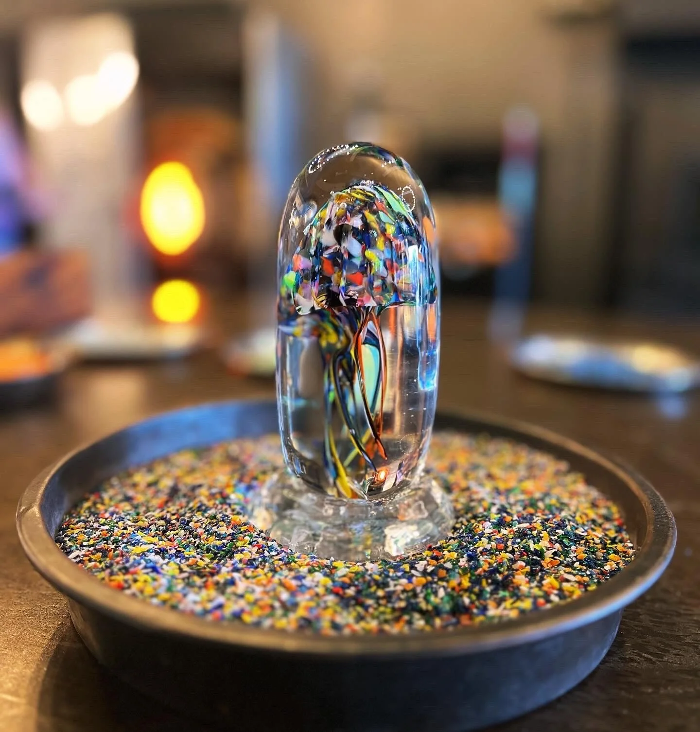 Glass sculpture resembling a jellyfish with colorful streaks inside, placed on a bed of multicolored glass frit in a black dish.