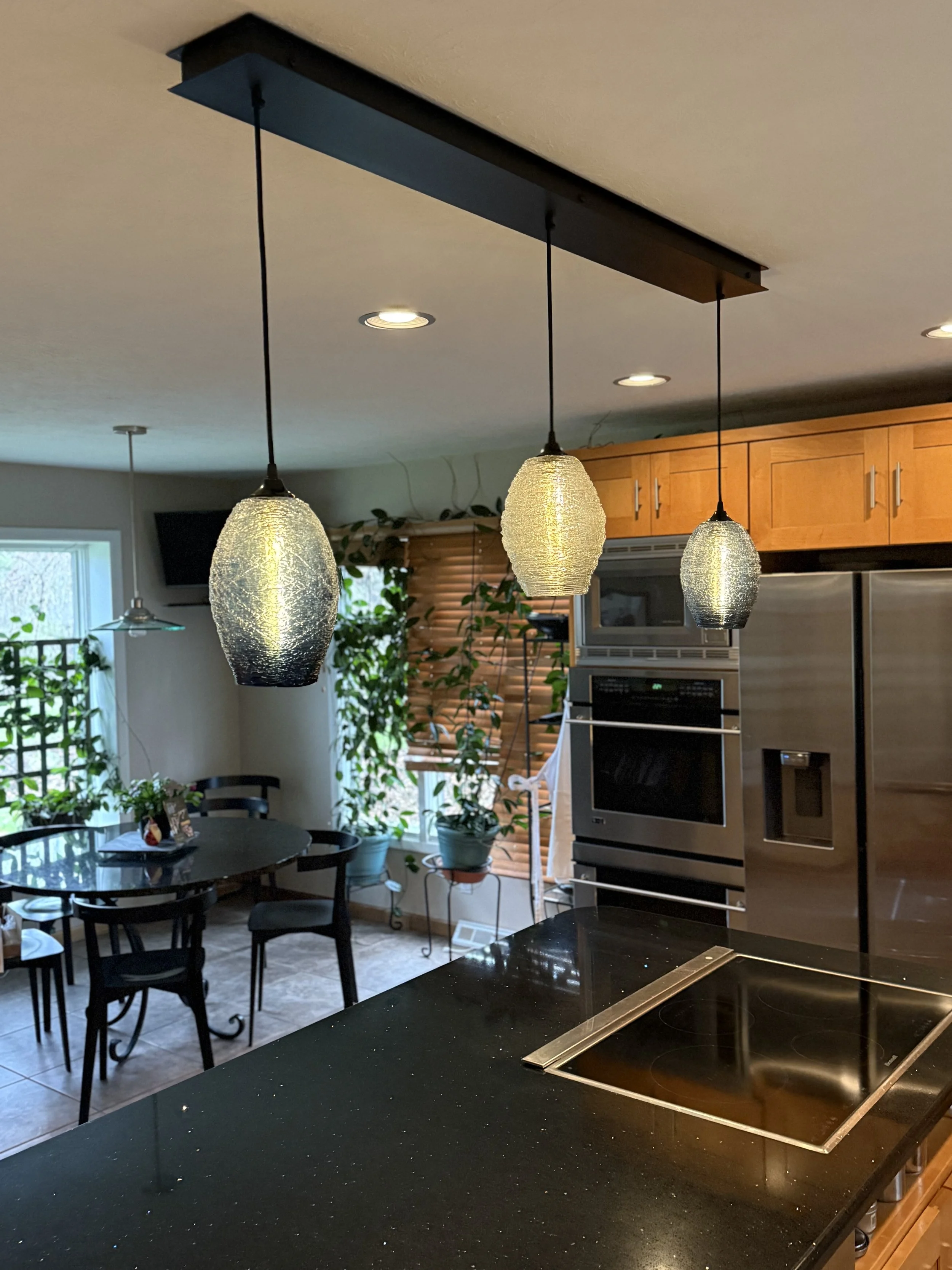 Interior view of a kitchen with three hanging pendant lights over a black granite counter, wooden cabinets, stainless steel appliances, a round dining table with black chairs, and large windows with blinds and plants outside.