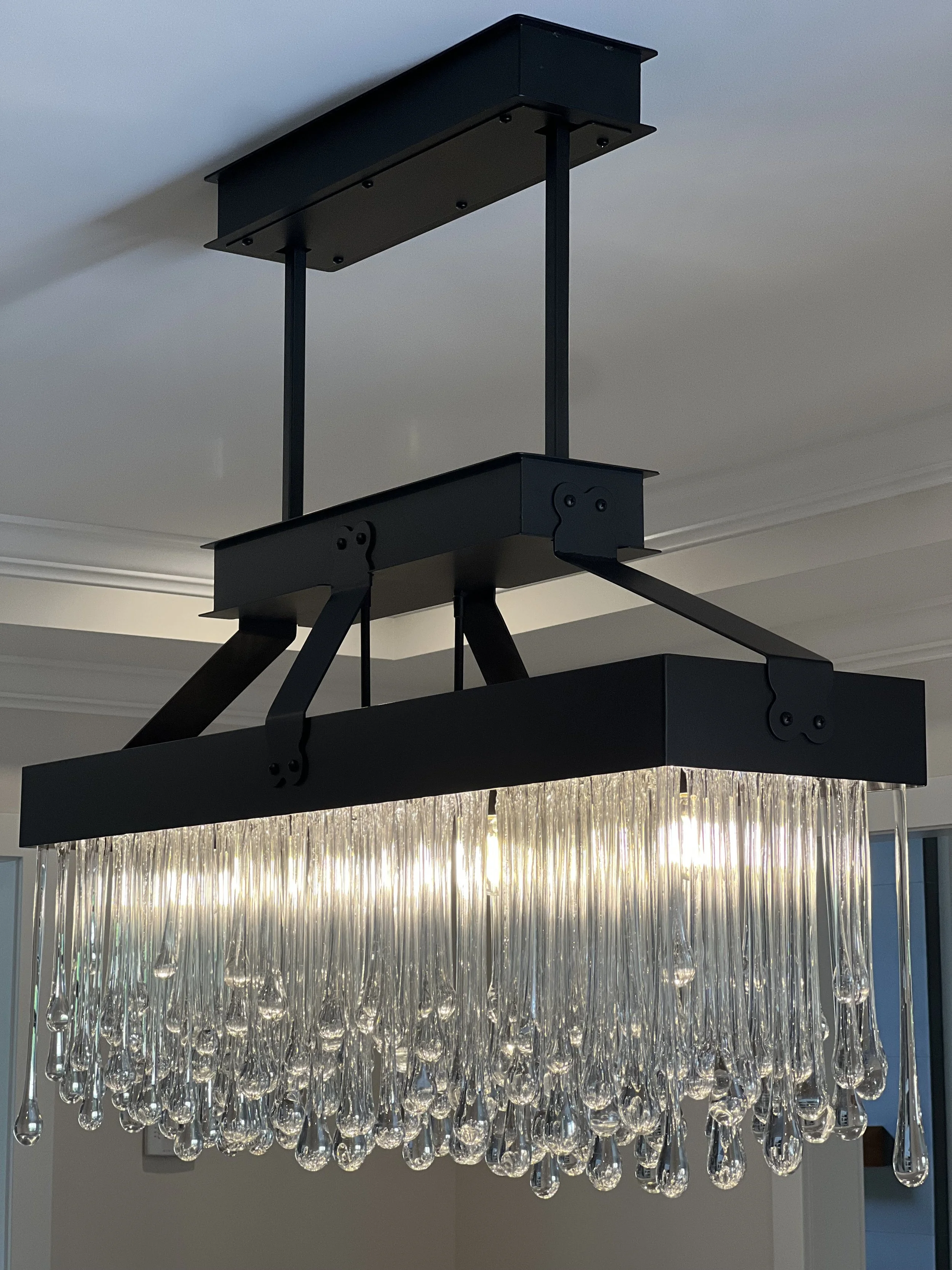 Modern chandelier with glass teardrop droplets hanging from a black metal frame, mounted on a ceiling in a room.