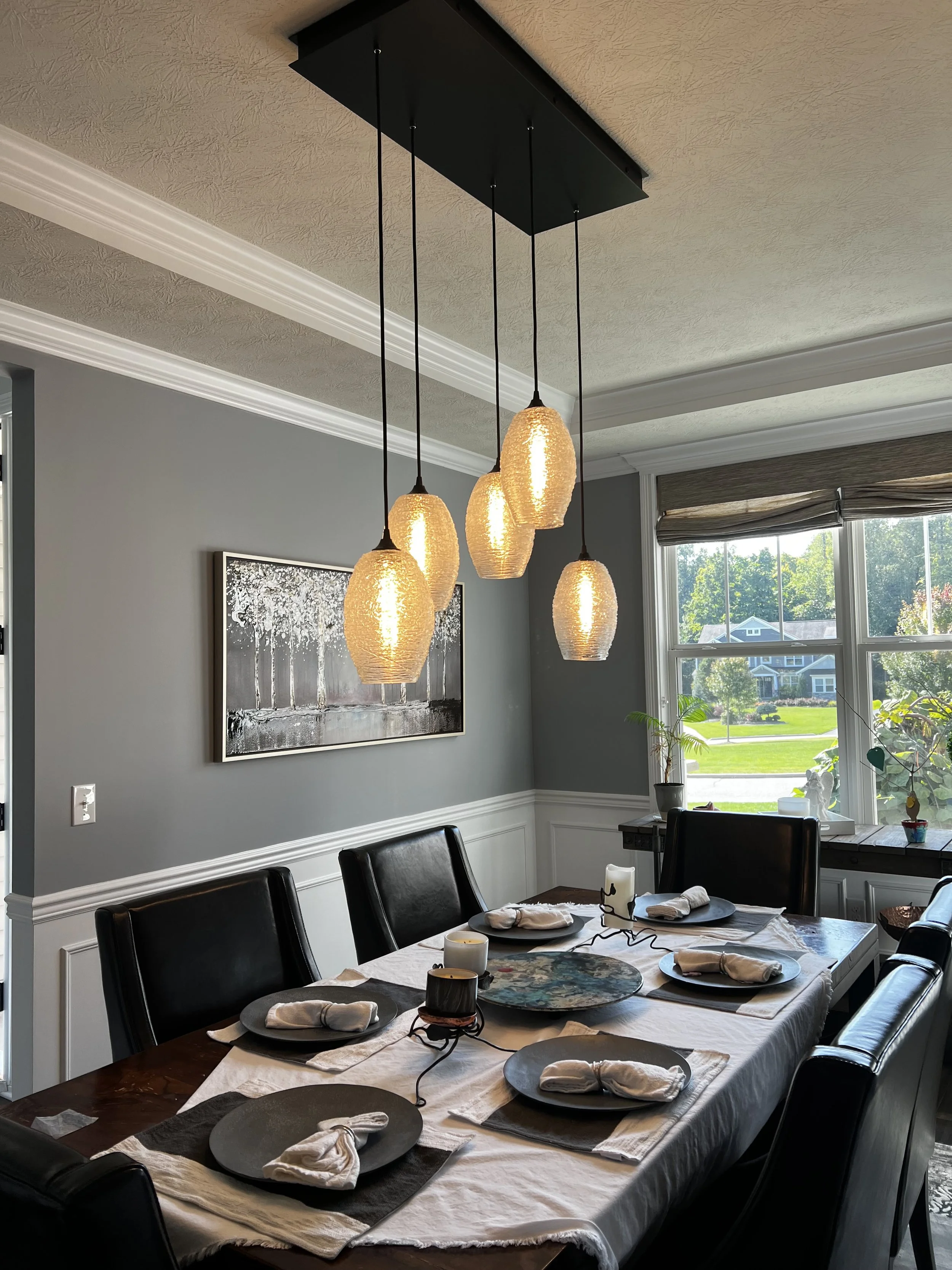 A dining room with a wooden table set for six, with dark plates, white napkins, and candles. Overhead, there are five hanging pendant lights with textured glass shades. The room has large windows with natural light and a view of a green lawn and hous