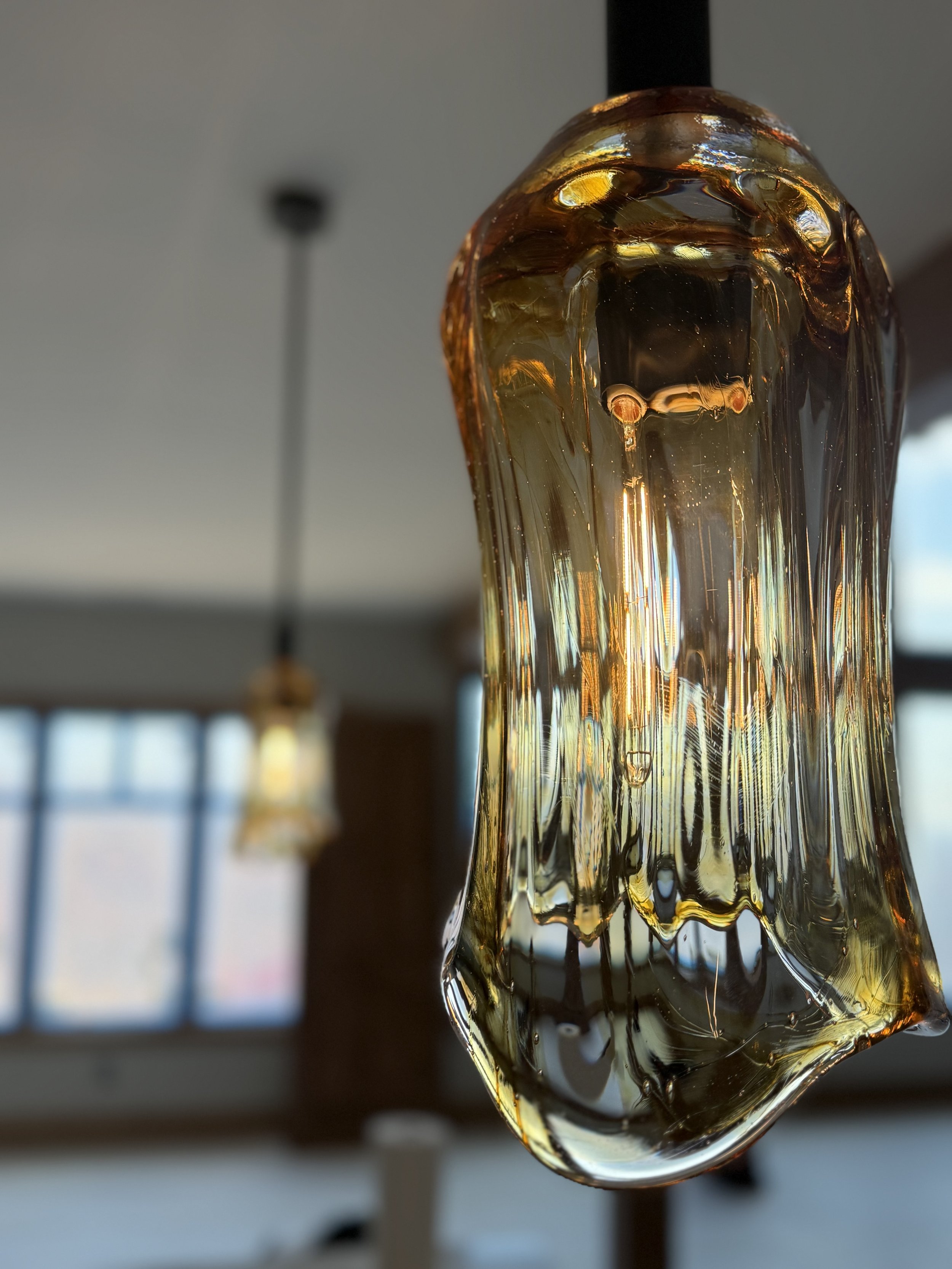 Close-up of a hanging glass art piece with amber and clear hues, reflecting light, with a blurred background of a room with windows and ceiling lights.