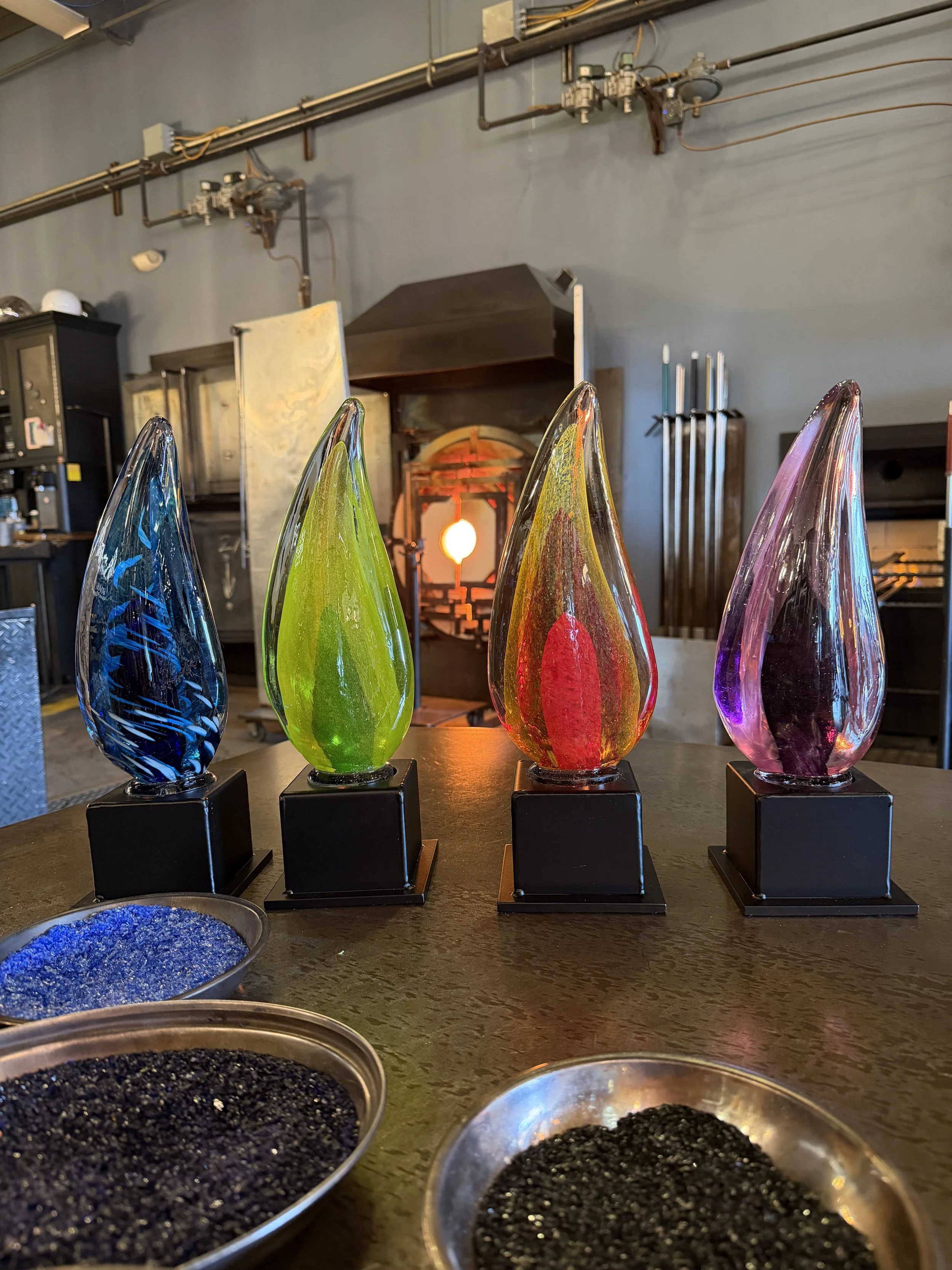 Four colorful glass art pieces in blue, green, red, and purple on black bases, placed on a wooden table in front of a furnace in a glass-blowing studio.