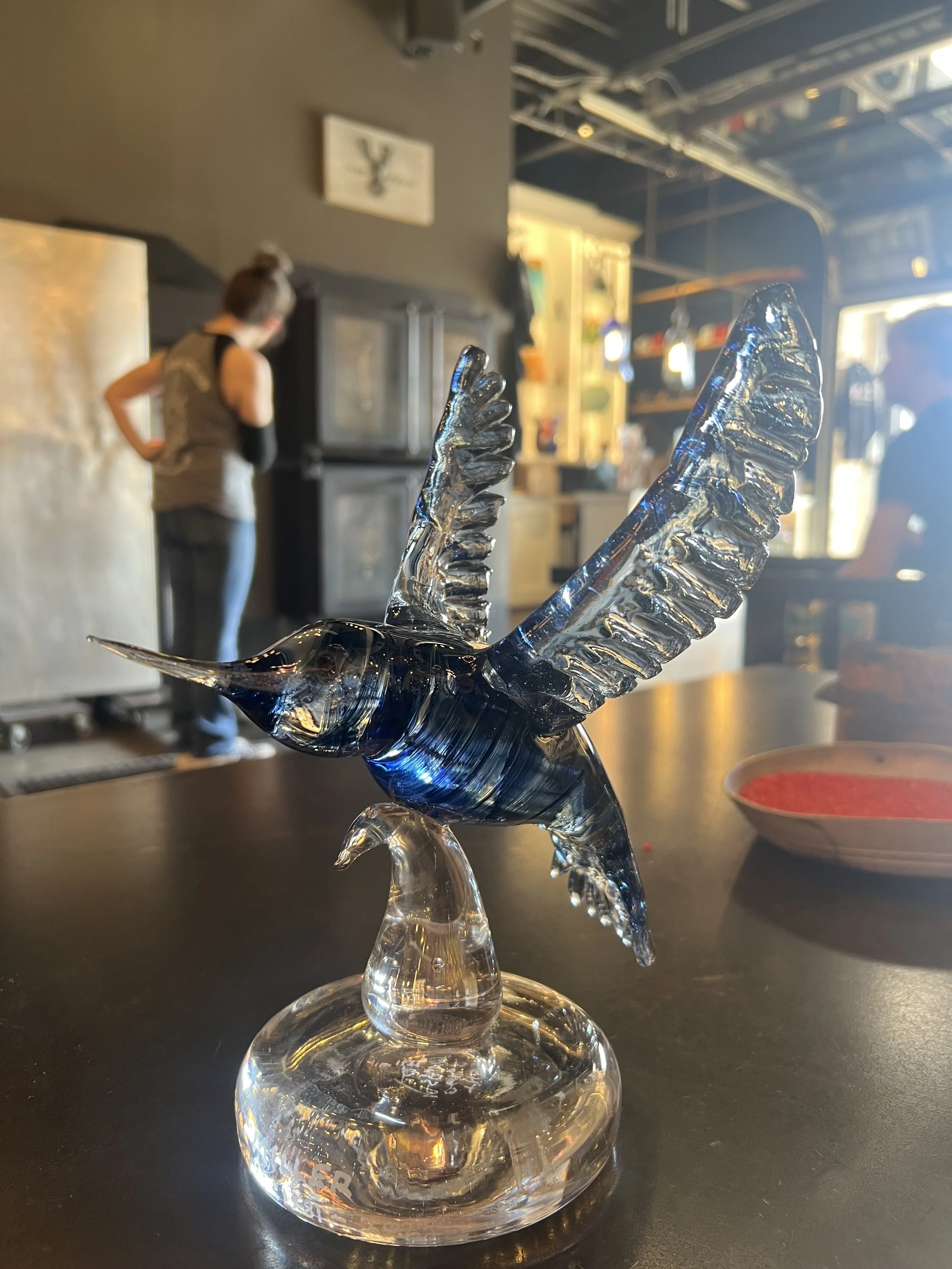 Glass sculpture of a bird with outstretched wings on a black table in a studio. Background includes a woman and a person, with some artwork on the walls.