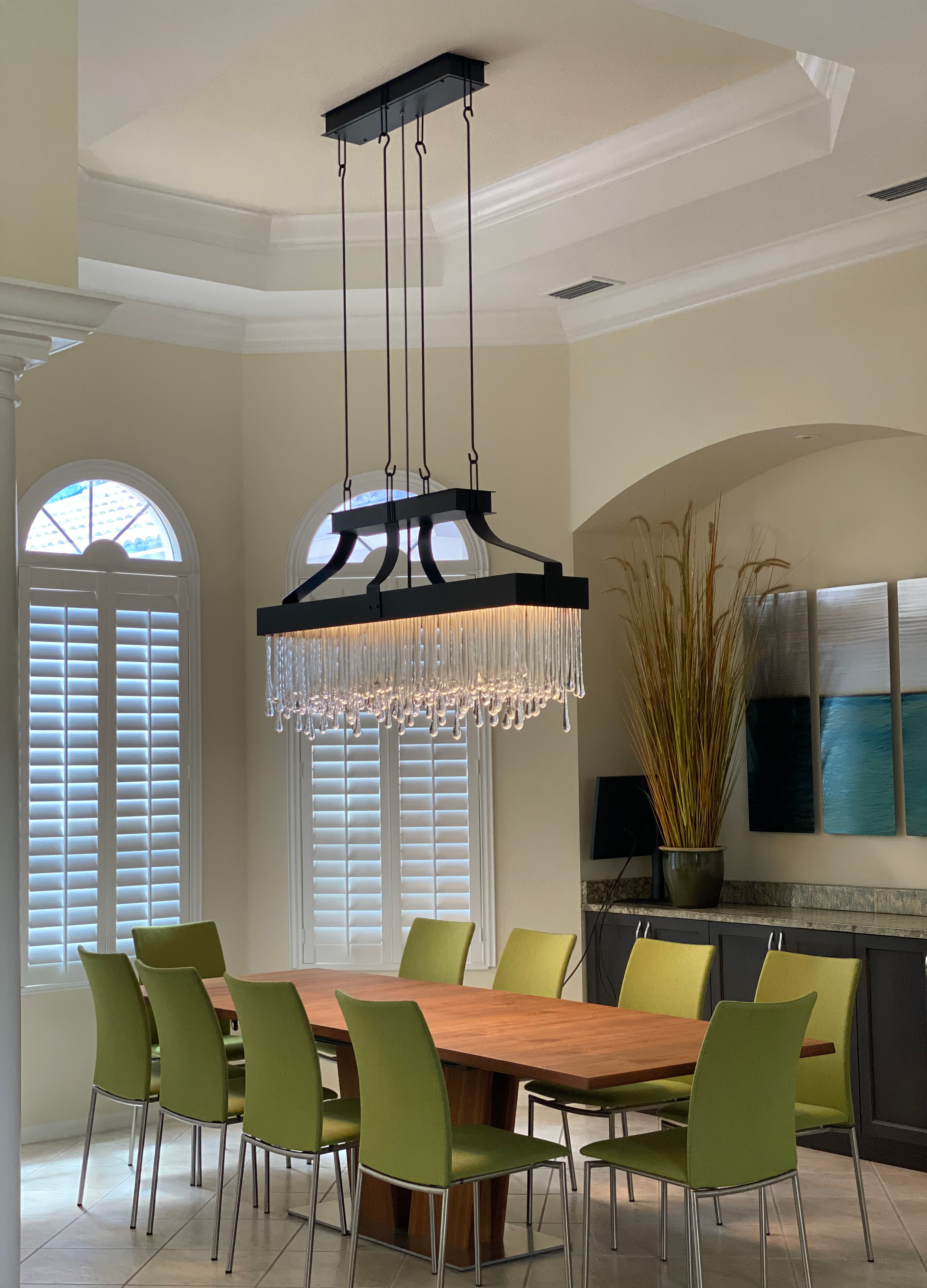 A dining room with a wooden table, ten green chairs, a modern chandelier with glass decorations, and windows with white shutters.