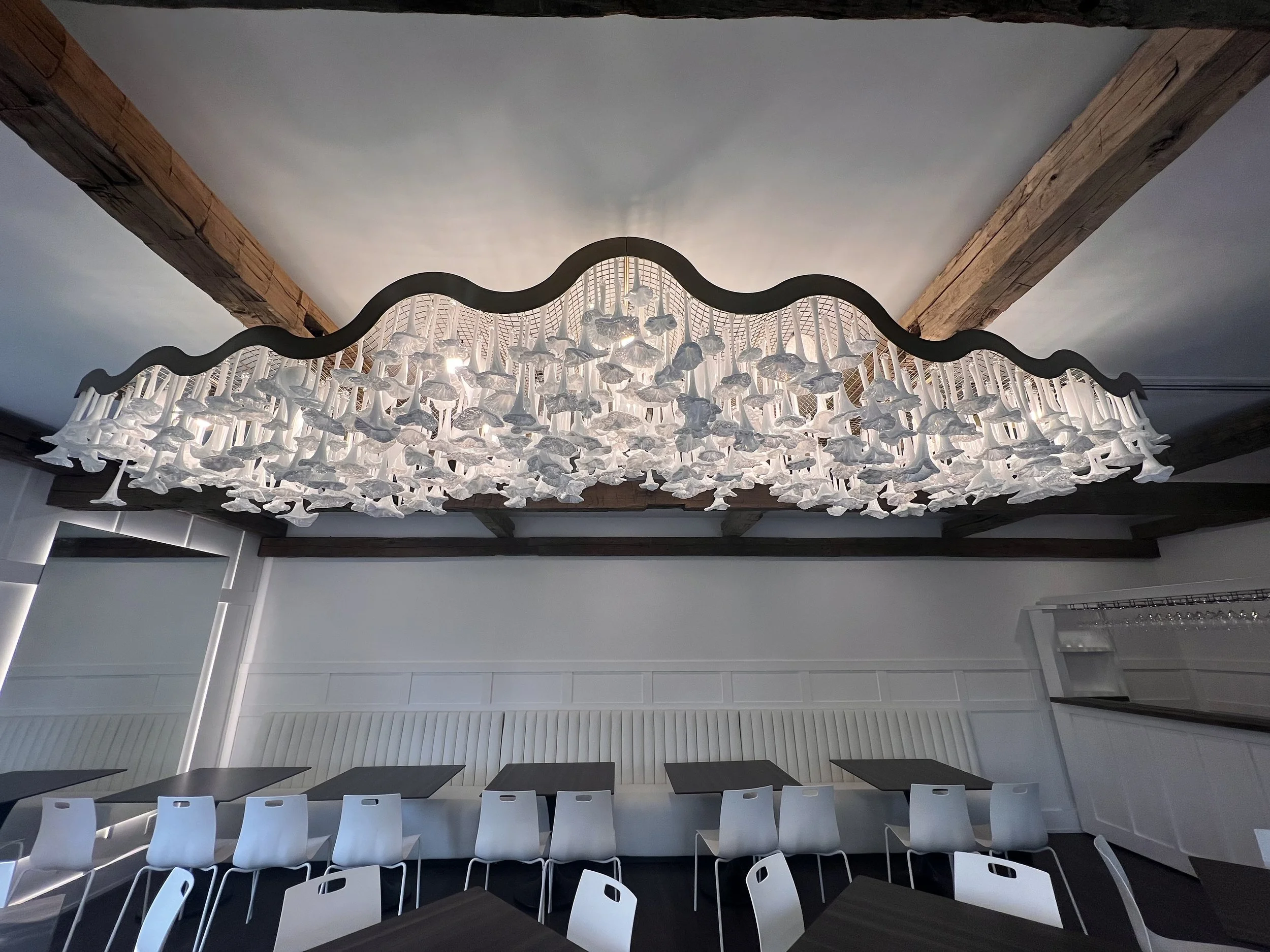 Modern chandelier made of hanging white fabric or paper swirls, mounted on a black wavy frame, hanging from a ceiling with wooden beams in a white dining area.