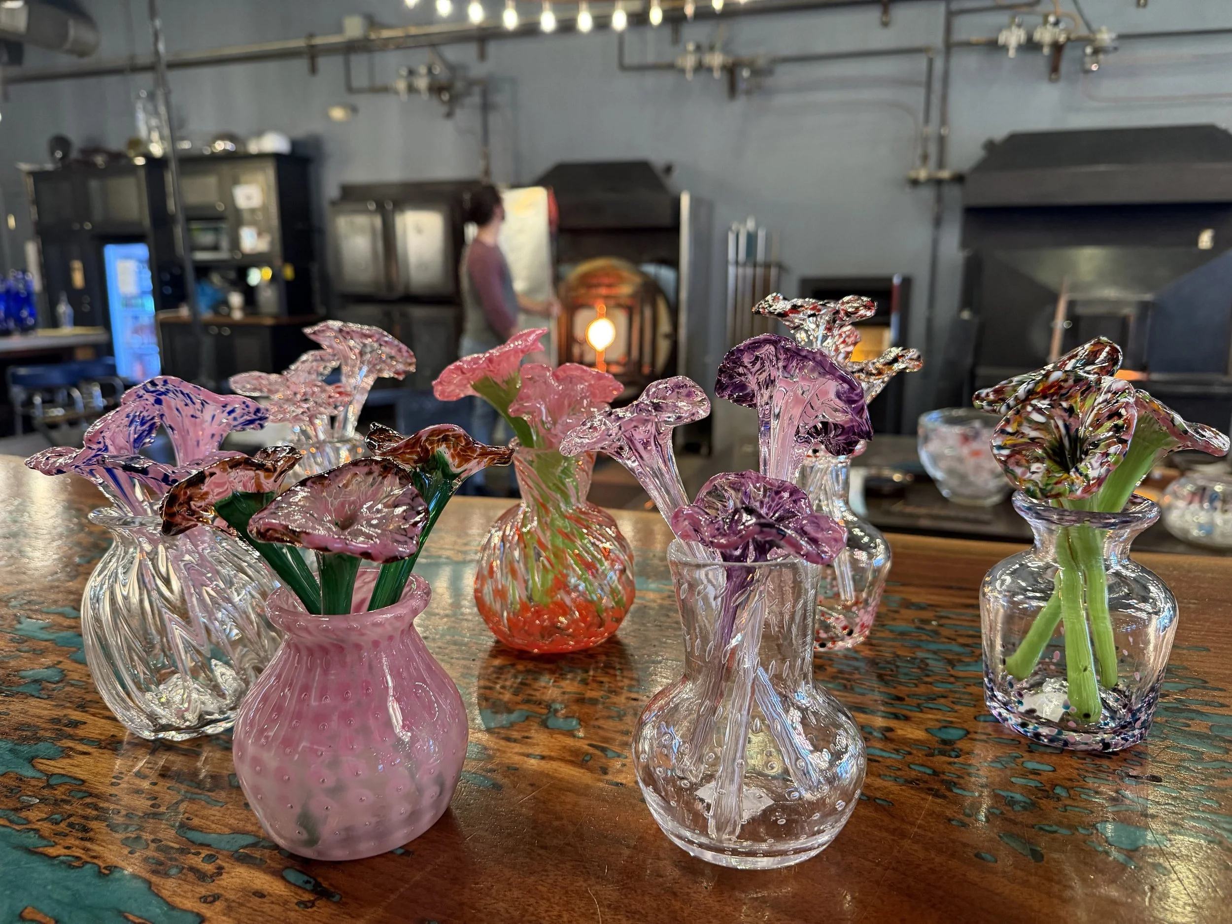 Colorful glass flower vases on a wooden table in a glass blowing studio with a person in the background.