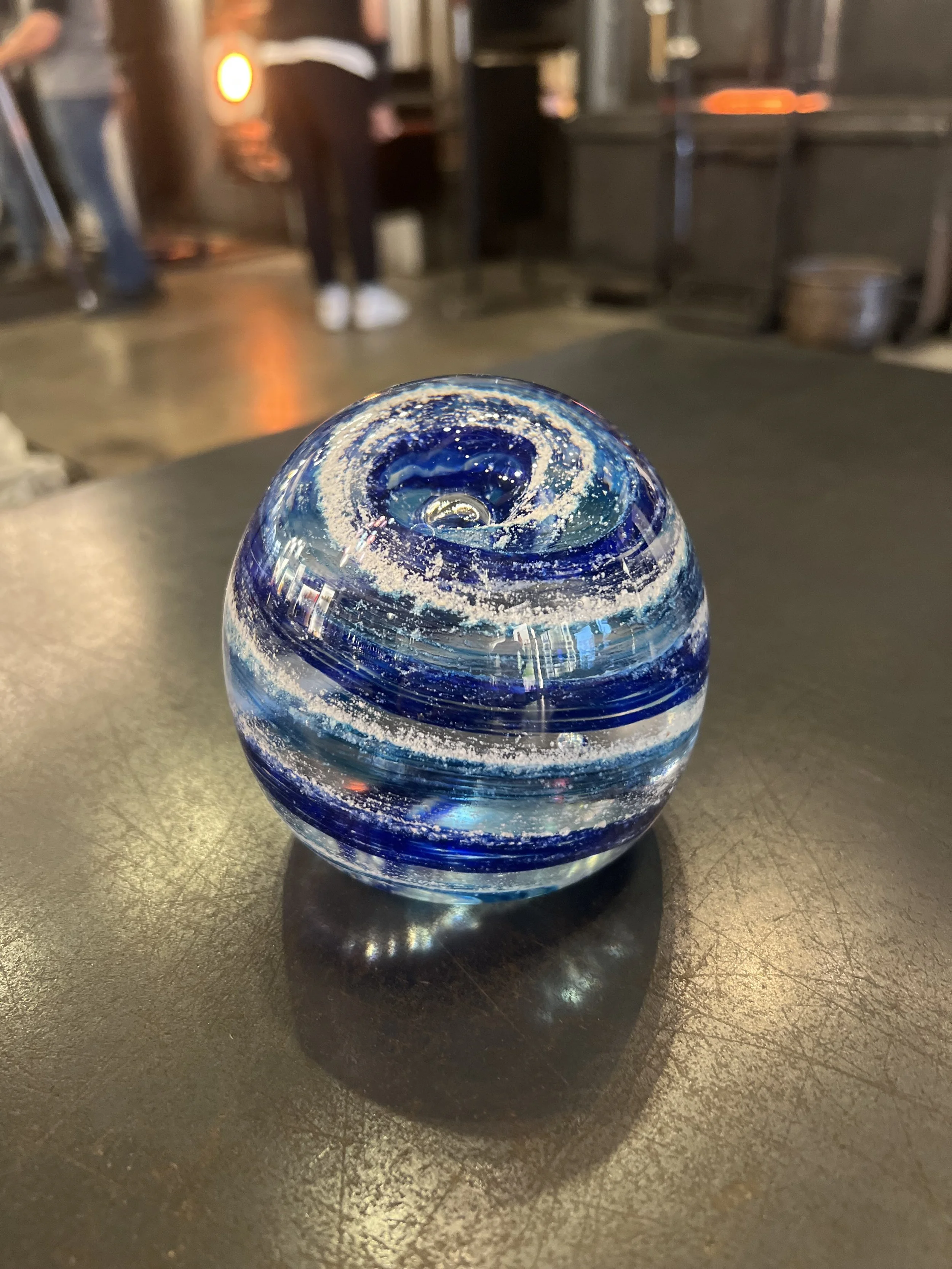A round, blue and white swirled marble with a glossy surface, placed on a metallic table in a dimly lit room with blurred background.