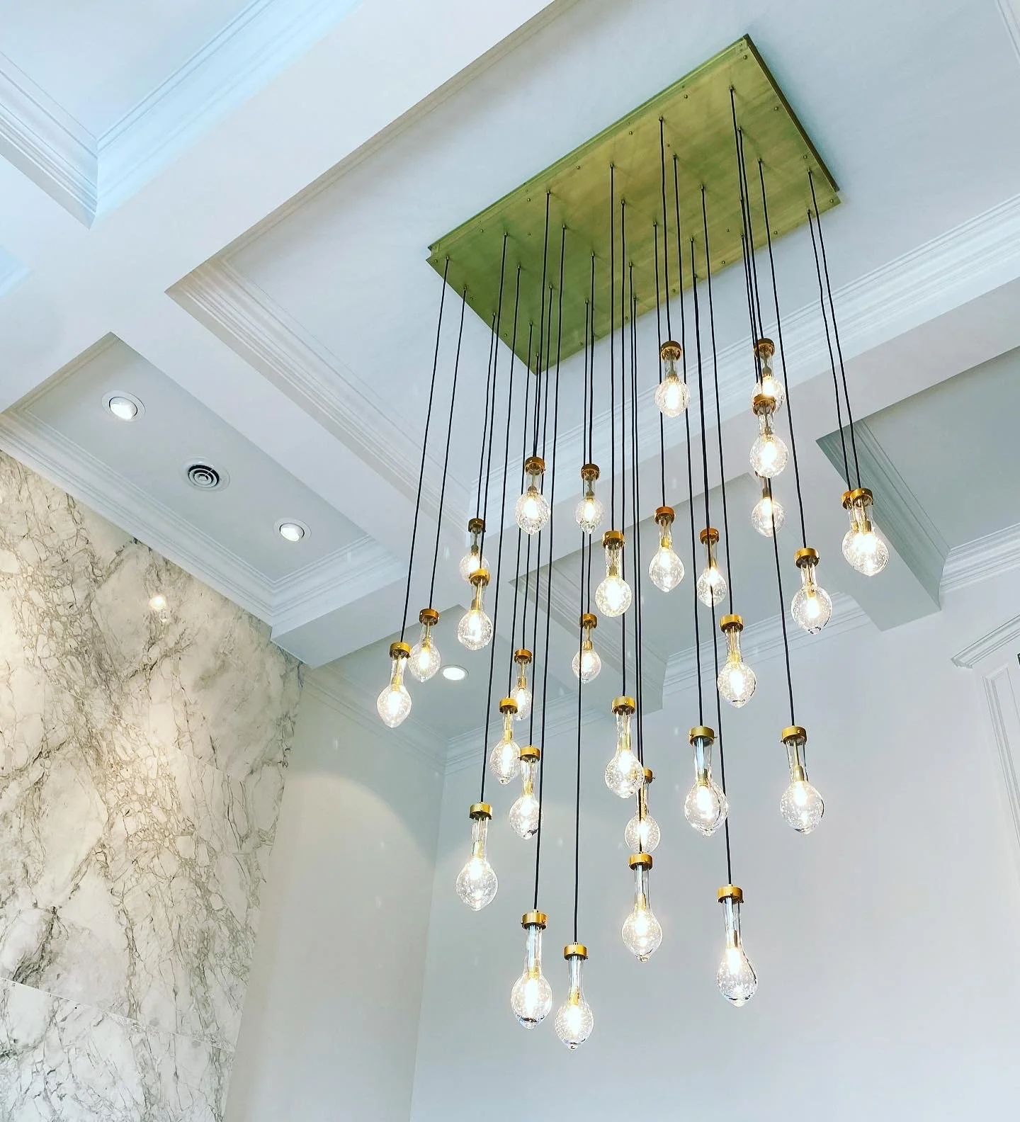A modern chandelier with multiple exposed light bulbs hanging from black cords on a green ceiling panel.