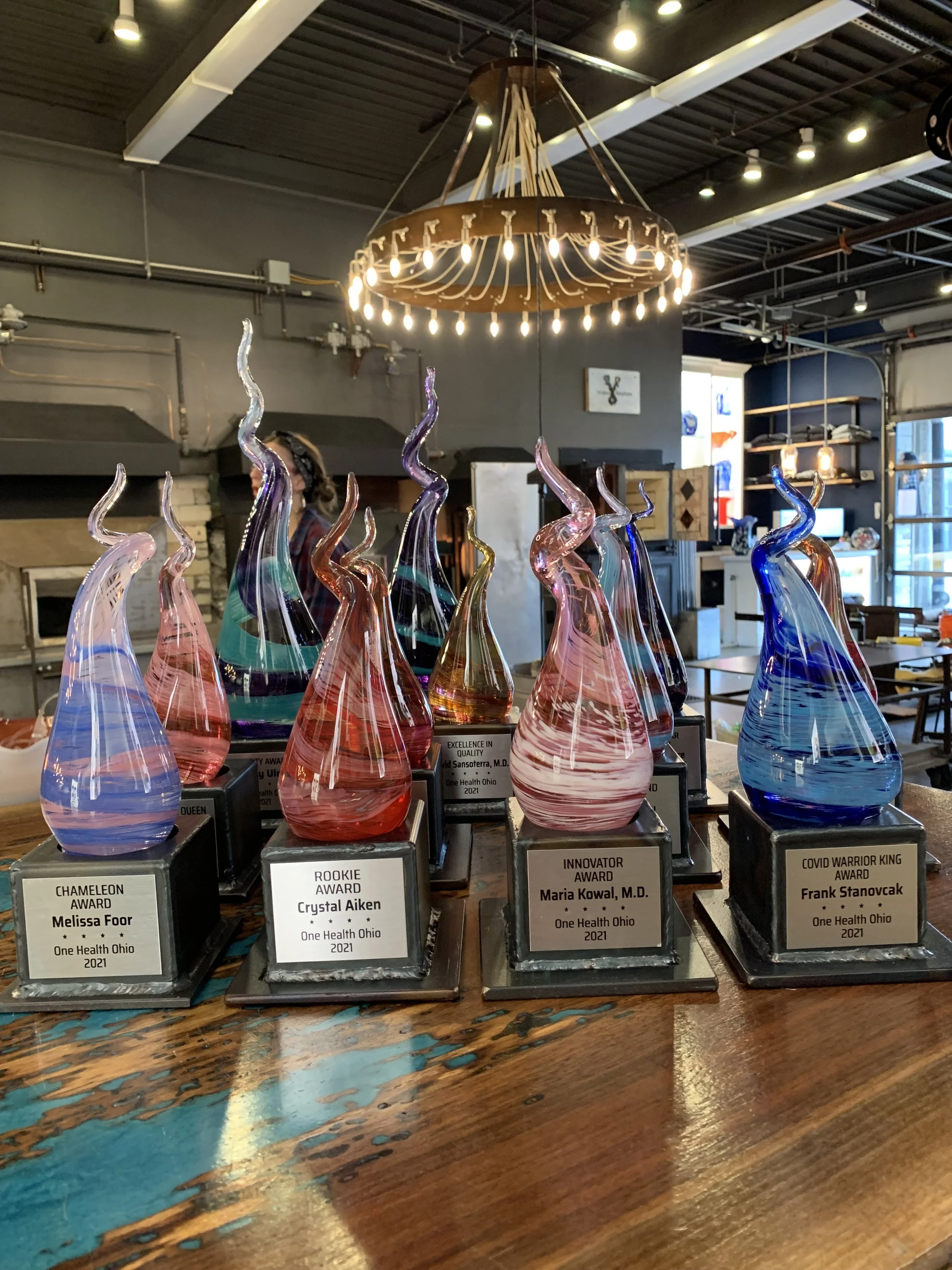 Colorful glass awards with spiral shapes on a wooden table, each labeled with different awards and recipients, inside a modern interior with a chandelier and shelves in the background.