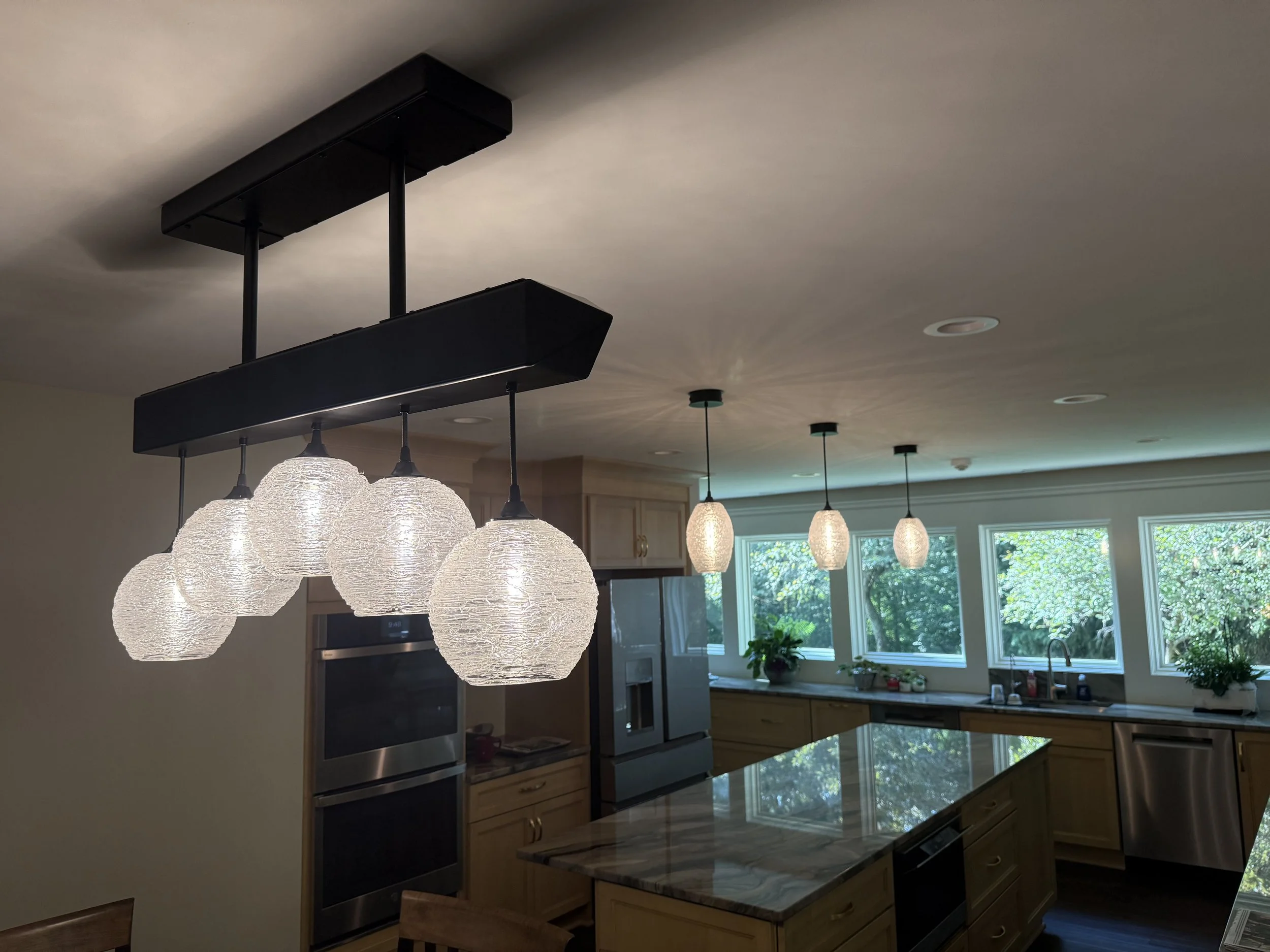 In the kitchen, a modern lighting fixture with multiple textured glass globe pendant lights hangs above a marble-topped island. The room features large windows with greenery outside, wooden cabinetry, and stainless steel appliances.