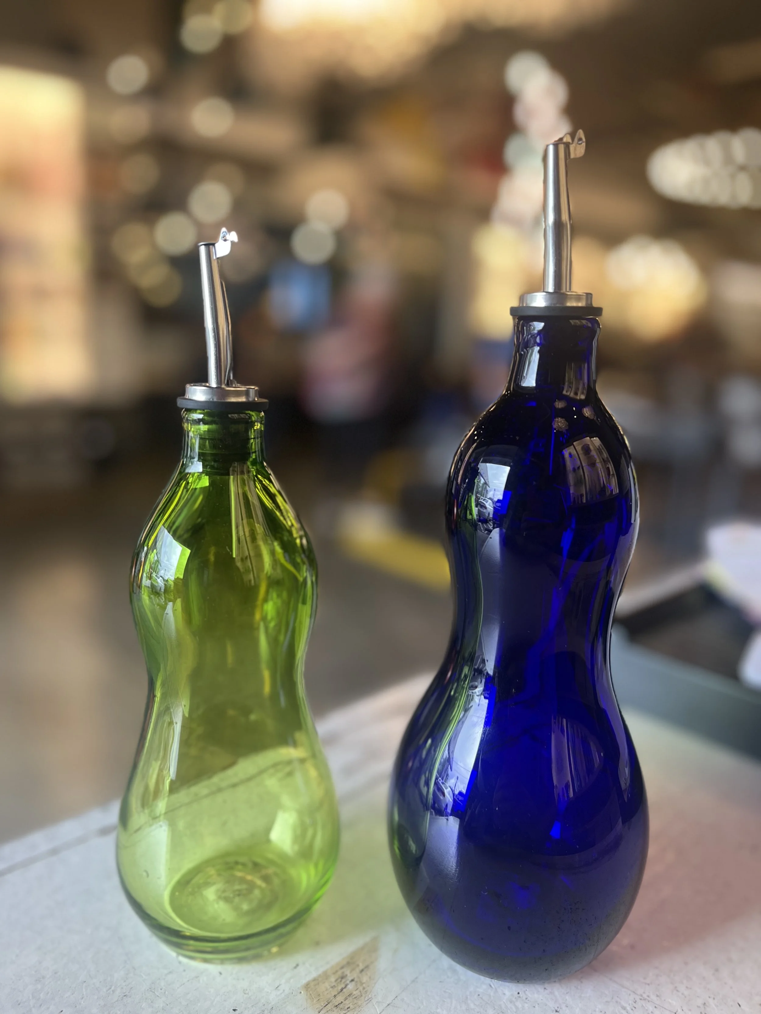 Two glass oil or vinegar bottles, one green and one blue, with metal pour spouts, on a countertop with a blurred store background.