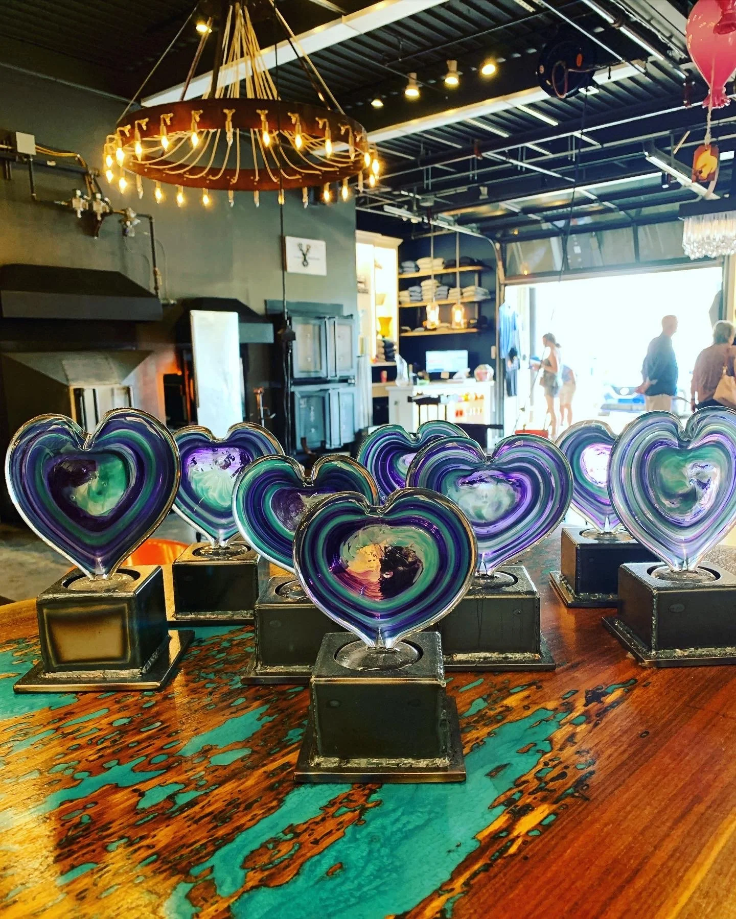 Colorful glass heart sculptures on display on a wooden table in a glass studio with people shopping in the background.