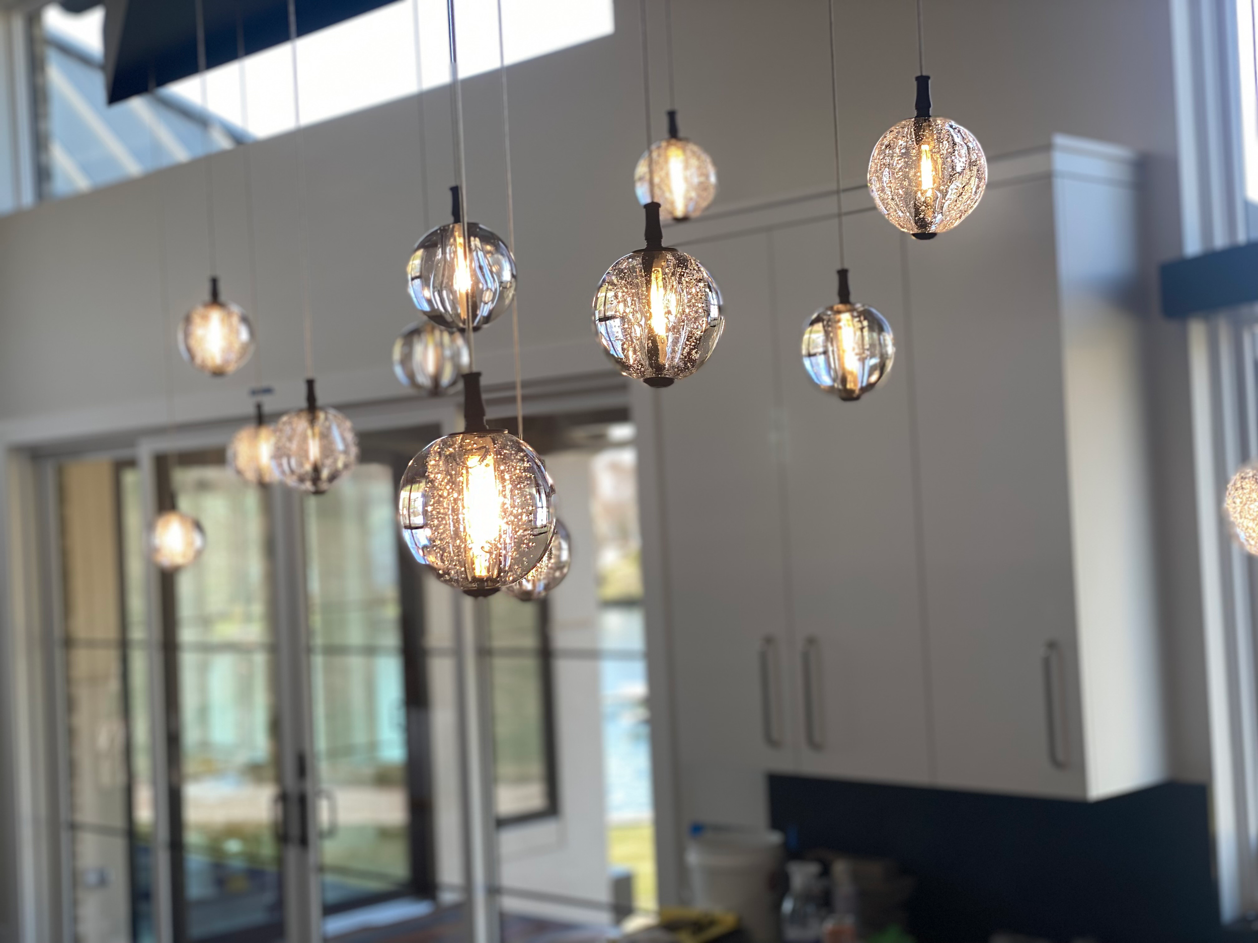 Multiple hanging pendant lights with reflective, textured glass globes in a modern indoor space with large windows and white cabinetry.