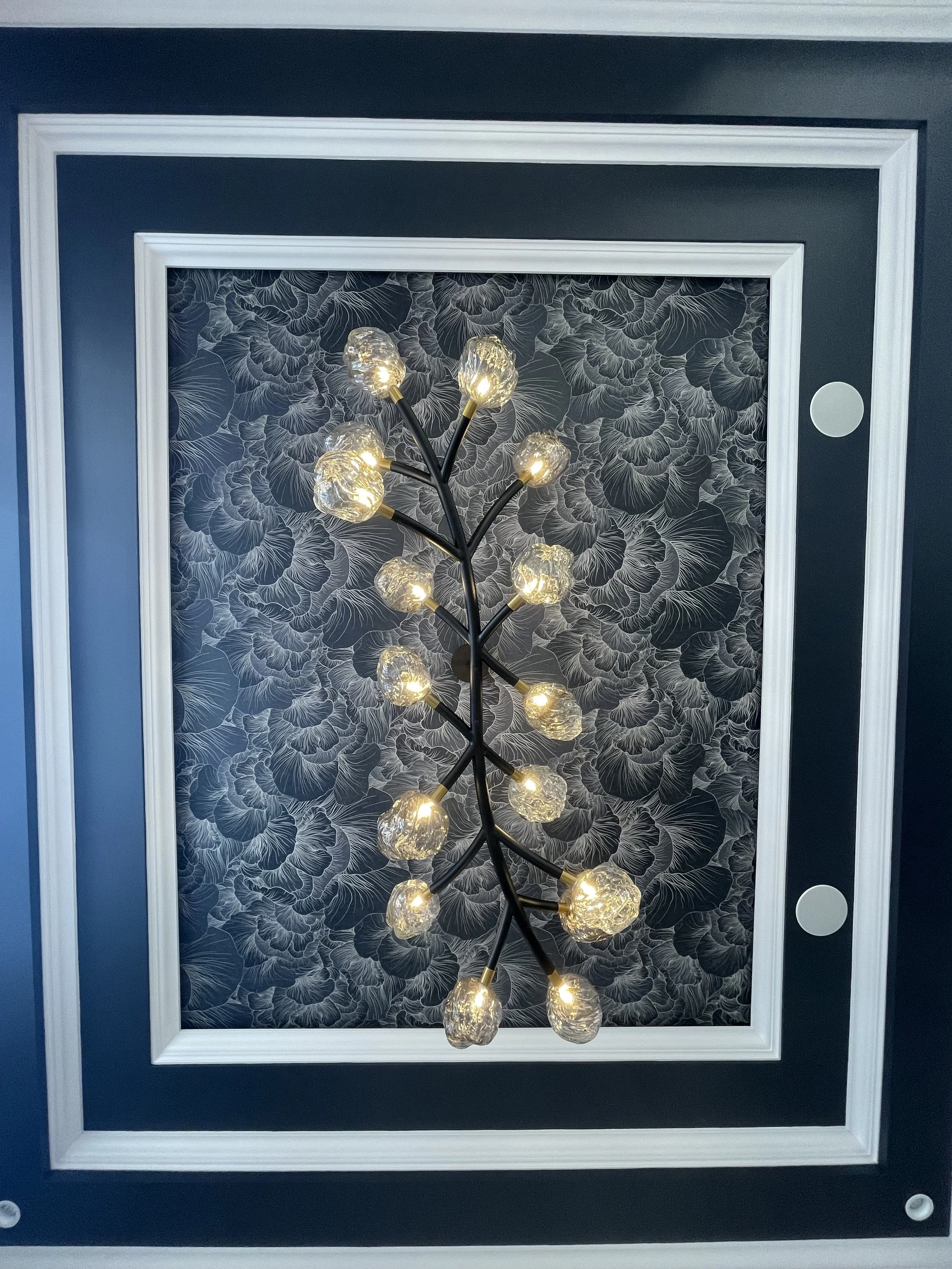 Modern ceiling light fixture with multiple glass globes resembling a branch, set against floral patterned black and white wallpaper in a ceiling with white and dark blue molding.
