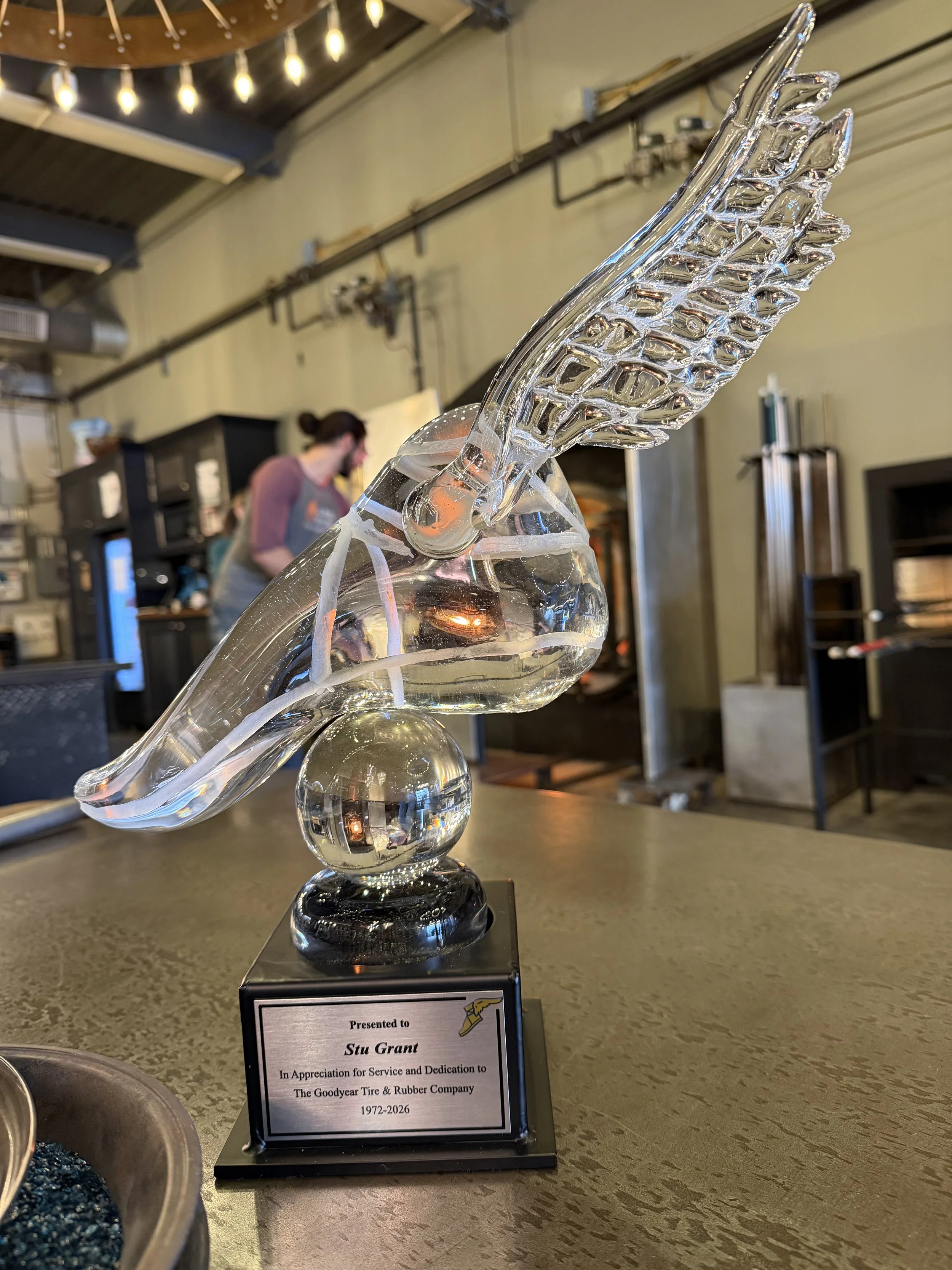 Glass trophy in the shape of a foot with wings, mounted on a black base with engraved plaque, in a studio. 