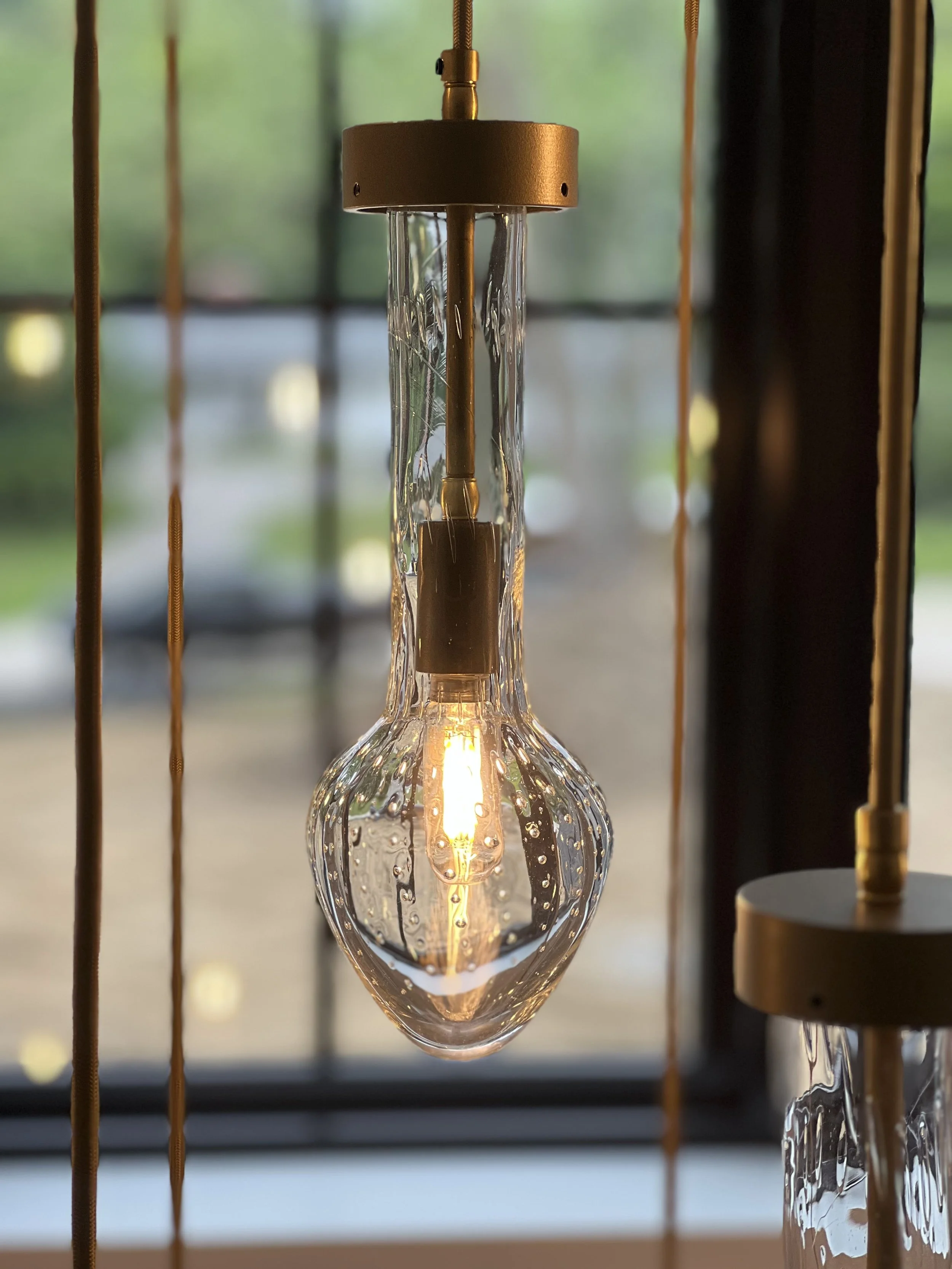 Close-up of a modern pendant light fixture with an exposed filament bulb hanging in front of a window with blurred greenery outside.