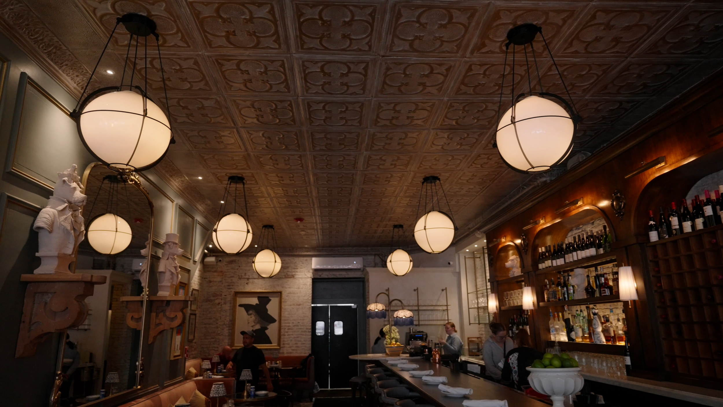 Interior of a cozy restaurant with ornate ceiling, hanging spherical lights, a bar area with shelves of wine bottles, and tables with patrons dining and a waitress serving.