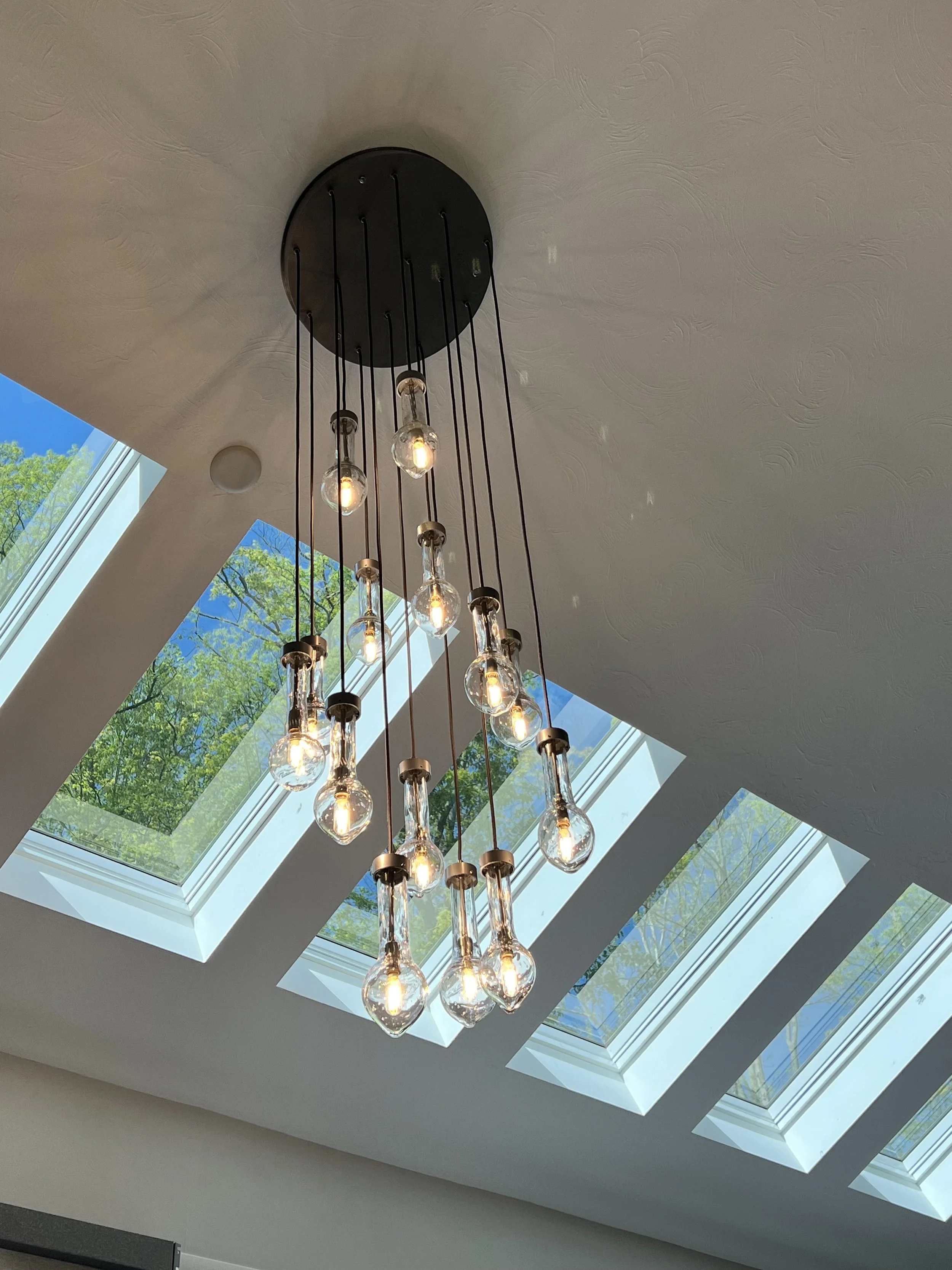Modern chandelier with multiple exposed light bulbs hanging from black cords, suspended from a ceiling with skylights showing green trees outside.