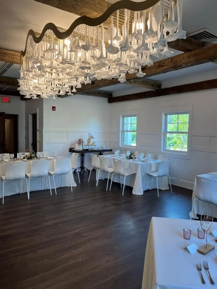 Elegant banquet hall with white walls, dark wood beams, and large windows. Decorated with white tablecloths, modern white chairs, and a unique hanging chandelier made of white cloth-like elements. The room has a polished wooden floor and is set up fo