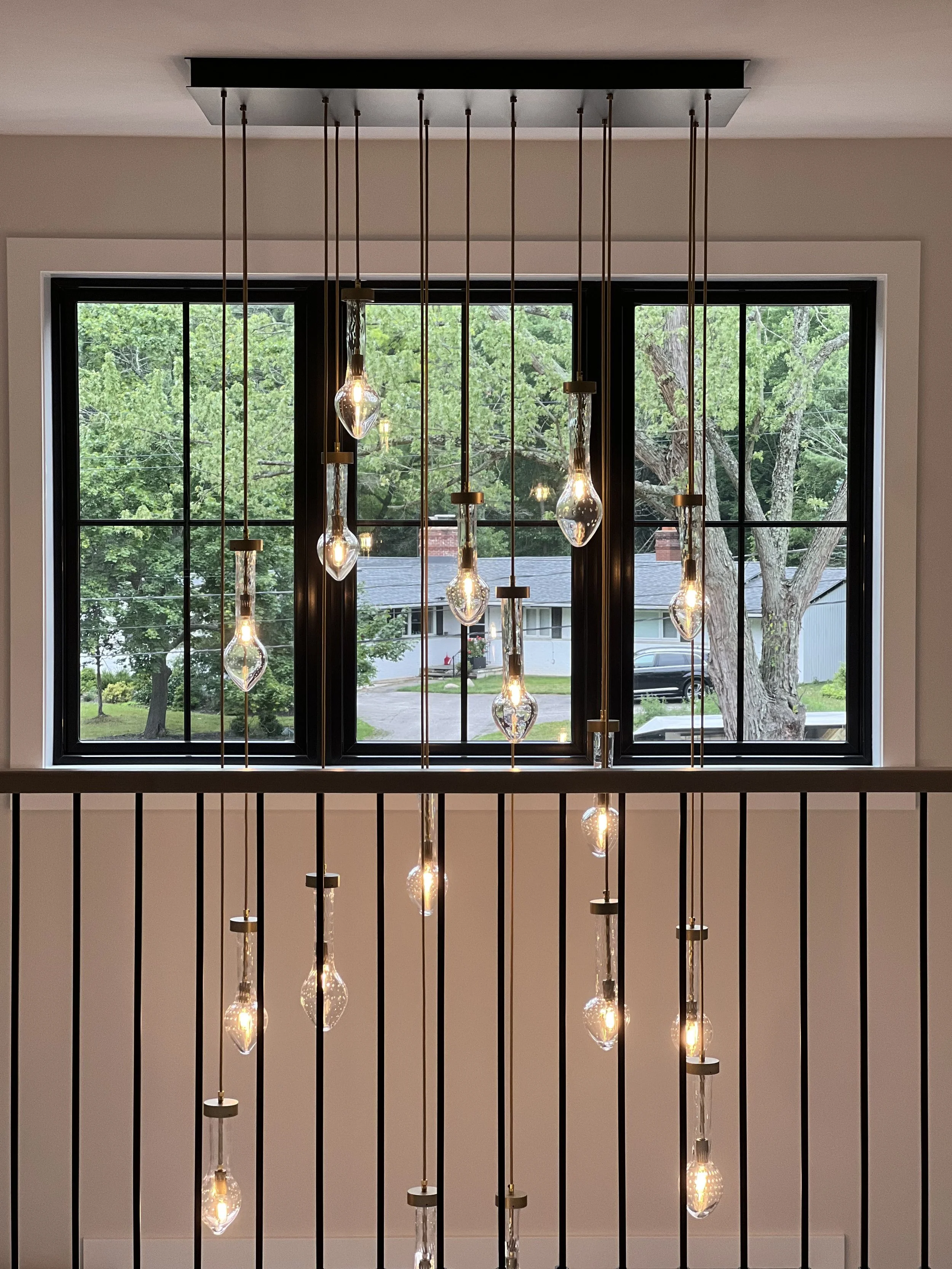Interior view of a modern chandelier with hanging glass light bulbs in front of a window with a view of trees and a neighborhood street.