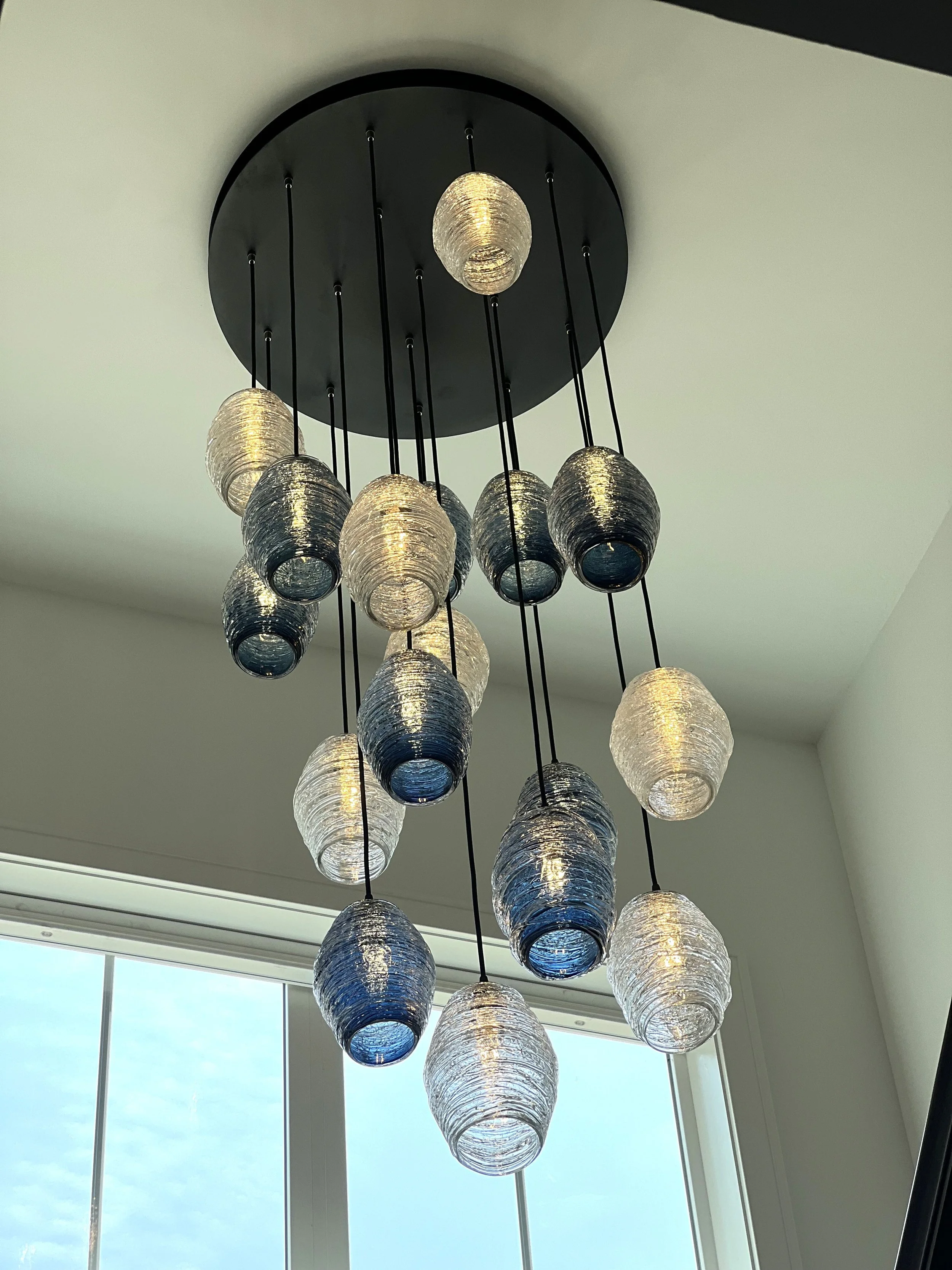 A modern chandelier with multiple hanging glass orbs in blue and white colors, suspended from a round ceiling mount, situated near a window with blinds.