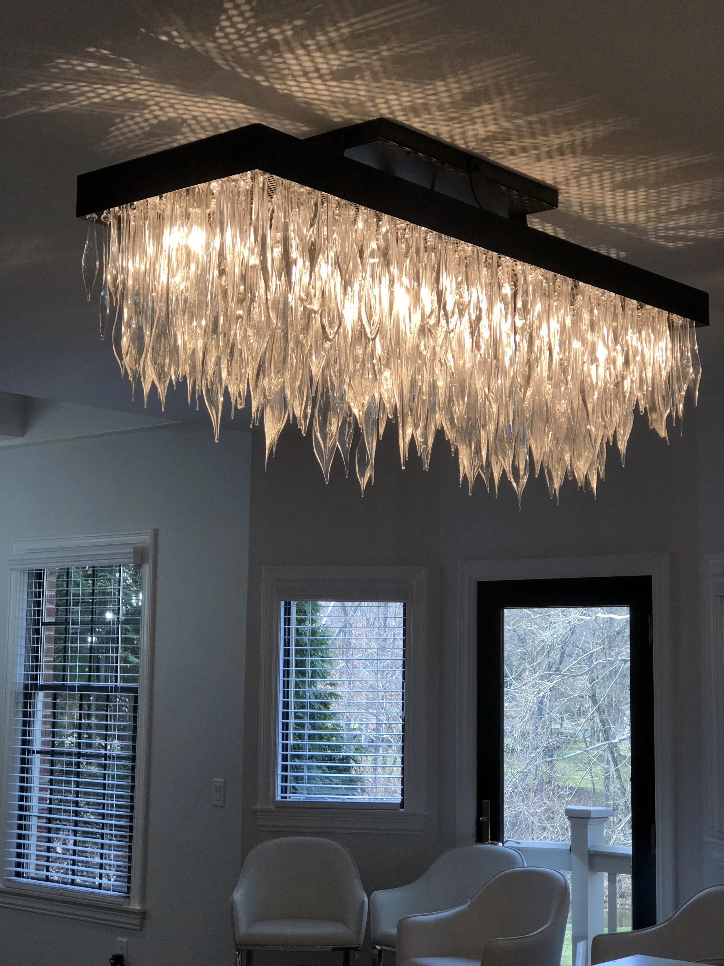 A modern chandelier with elongated glass droplets hanging from a rectangular black frame, casting intricate light patterns on the ceiling in a living room with white walls, white chairs, and a glass door opening to an outdoor view.