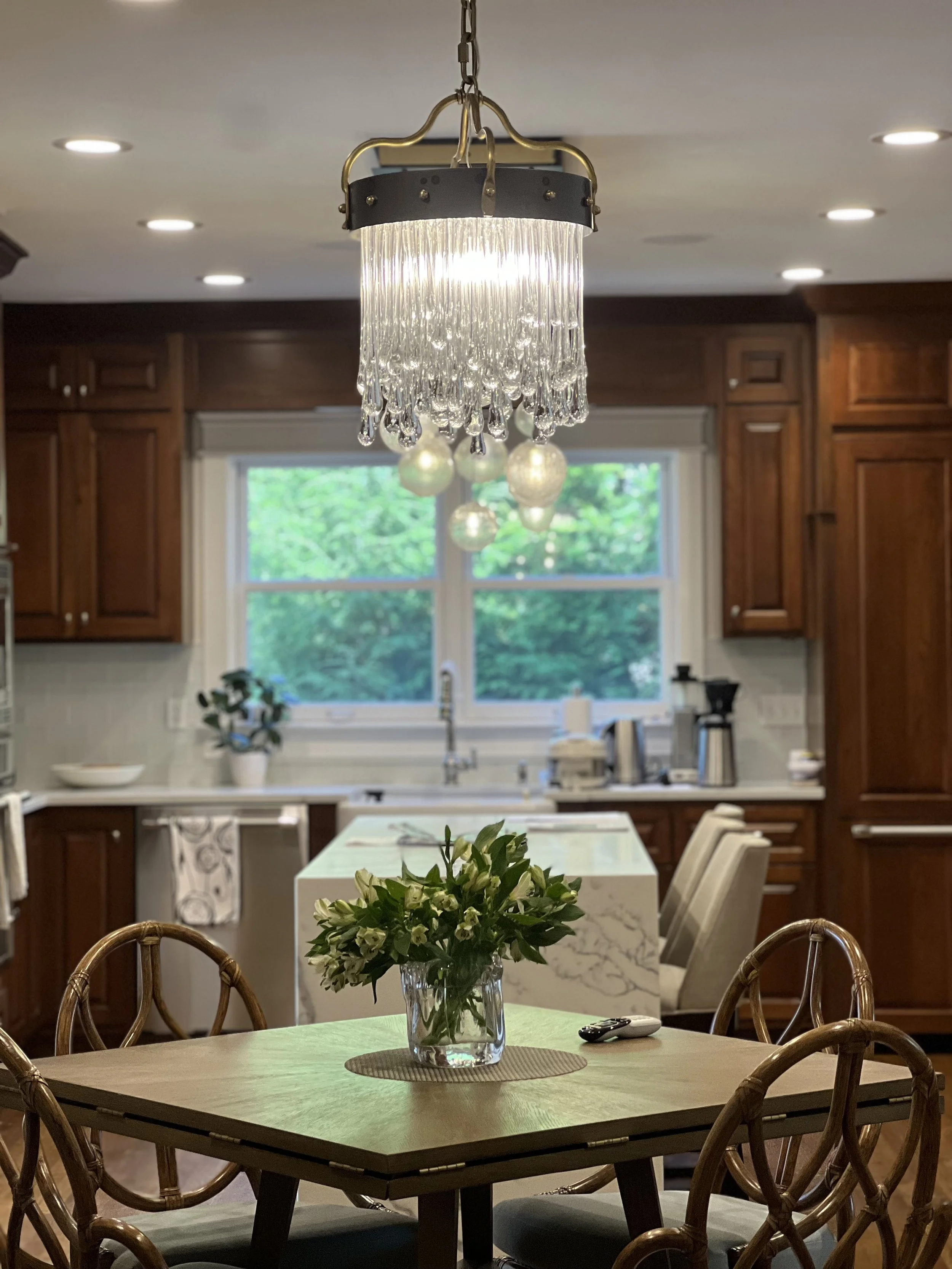 A kitchen with a round wooden table adorned with a glass vase holding a bouquet of white flowers. A chandelier with hanging glass droplets is above the table. In the background, wooden cabinets, a window with green trees outside, and a kitchen island