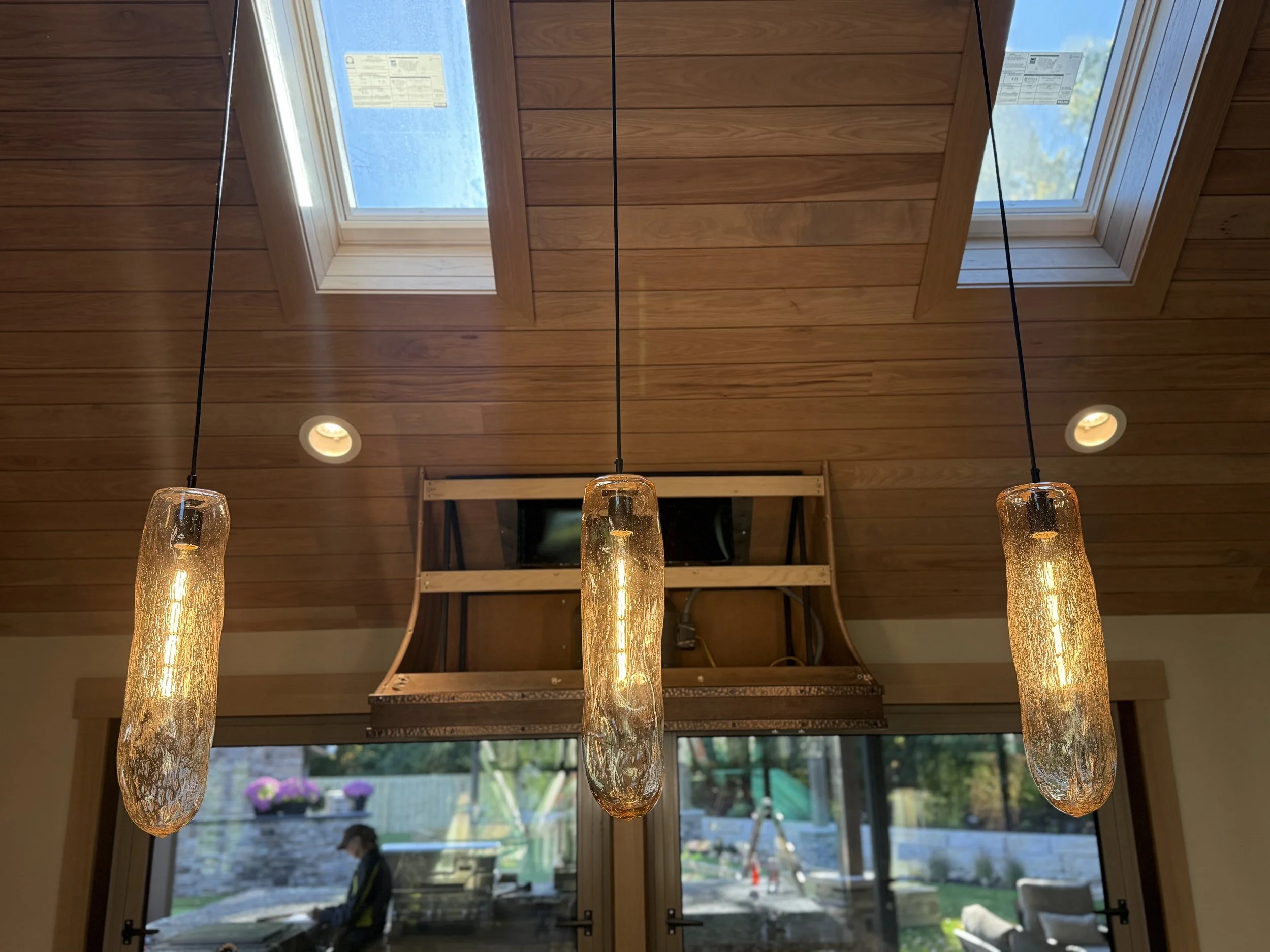 Modern pendant lights with elongated glass shades hanging from a wooden ceiling with skylights, overlooking a sunlit outdoor patio.