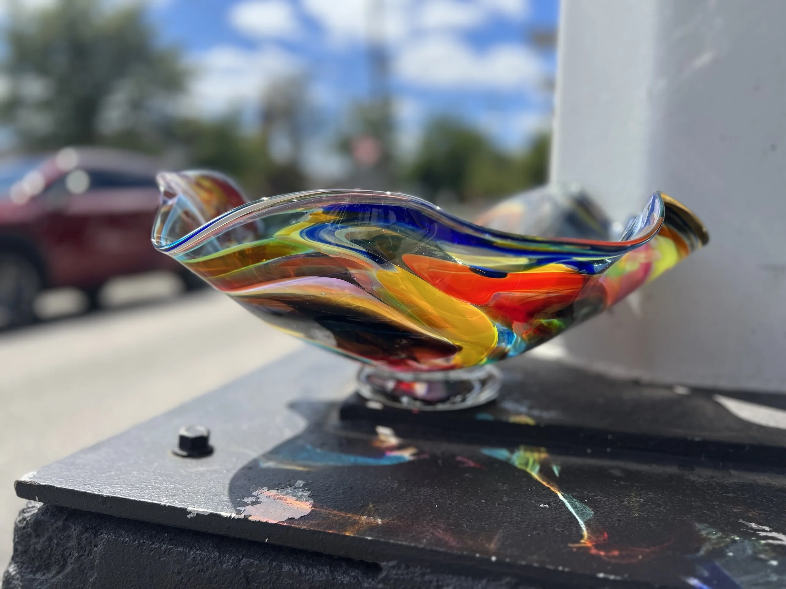 Colorful hand-blown glass sculpture with swirls of red, yellow, blue, and black, placed outdoors on a black surface with a blurred background.
