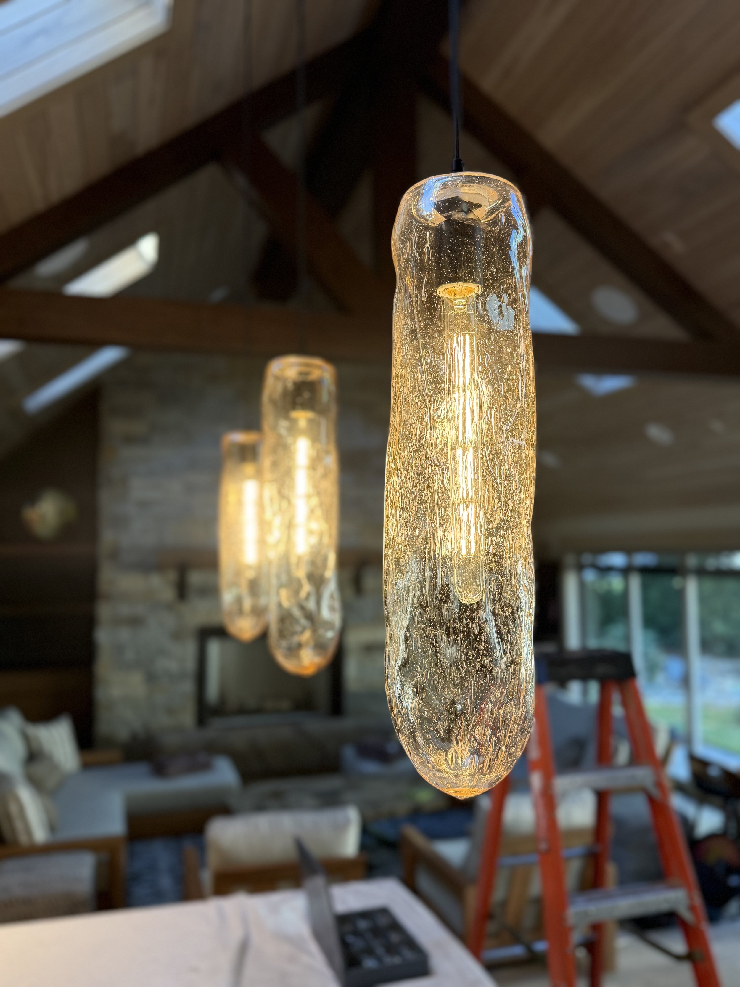 Close-up of three hanging pendant lights made of textured, elongated glass with warm glowing filament bulbs inside, in a cozy living room with wooden ceiling, stone fireplace, and large windows.