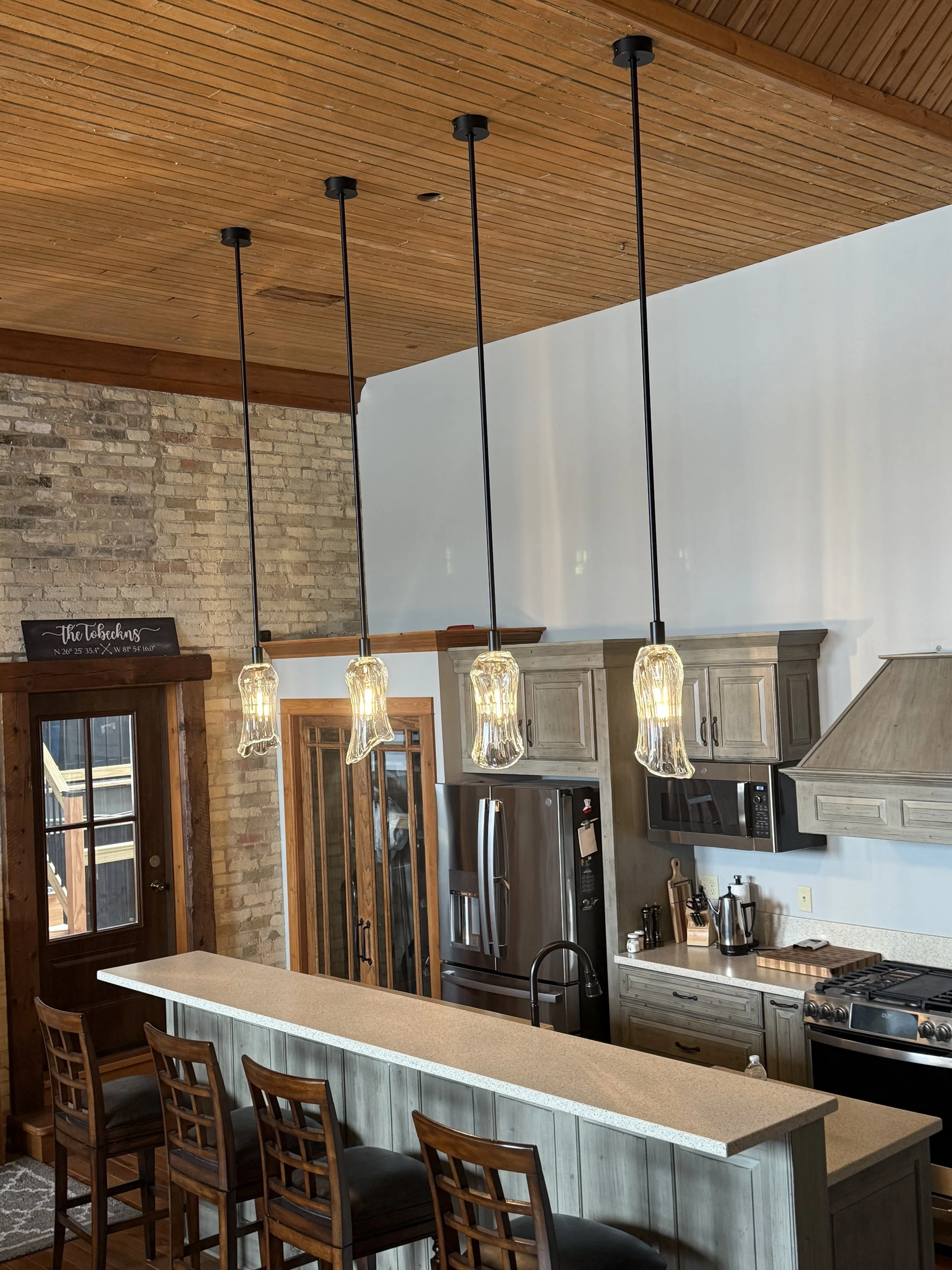 A modern kitchen with four pendant lights hanging above a beige kitchen island, stainless steel refrigerator, microwave, stove, and wooden cabinets, with a brick wall and wooden ceiling.