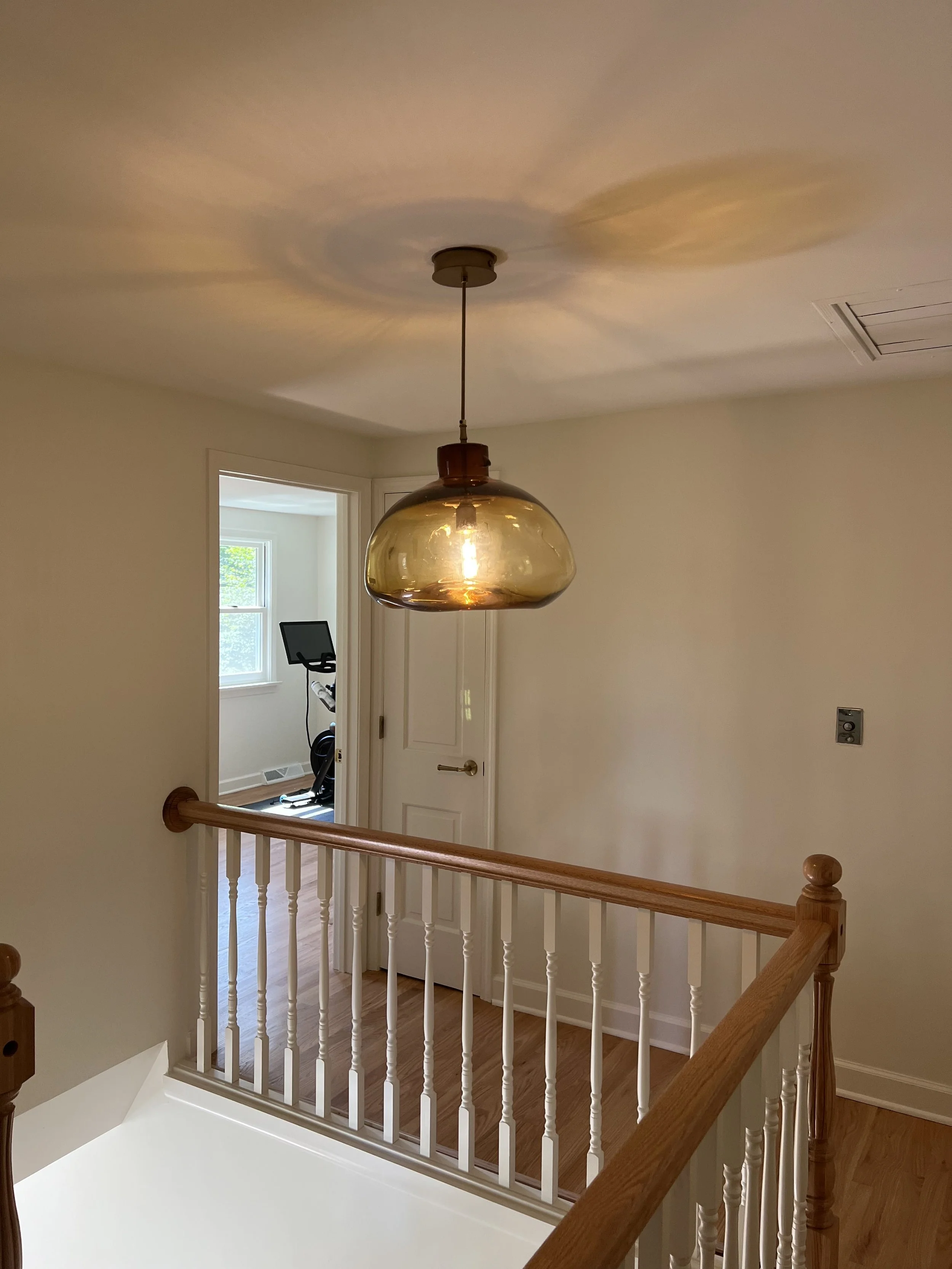 A ceiling light with a amber-colored glass shade hanging above wood handrail and white spindles, overlooking a hallway with a door and a room with exercise equipment in the background.
