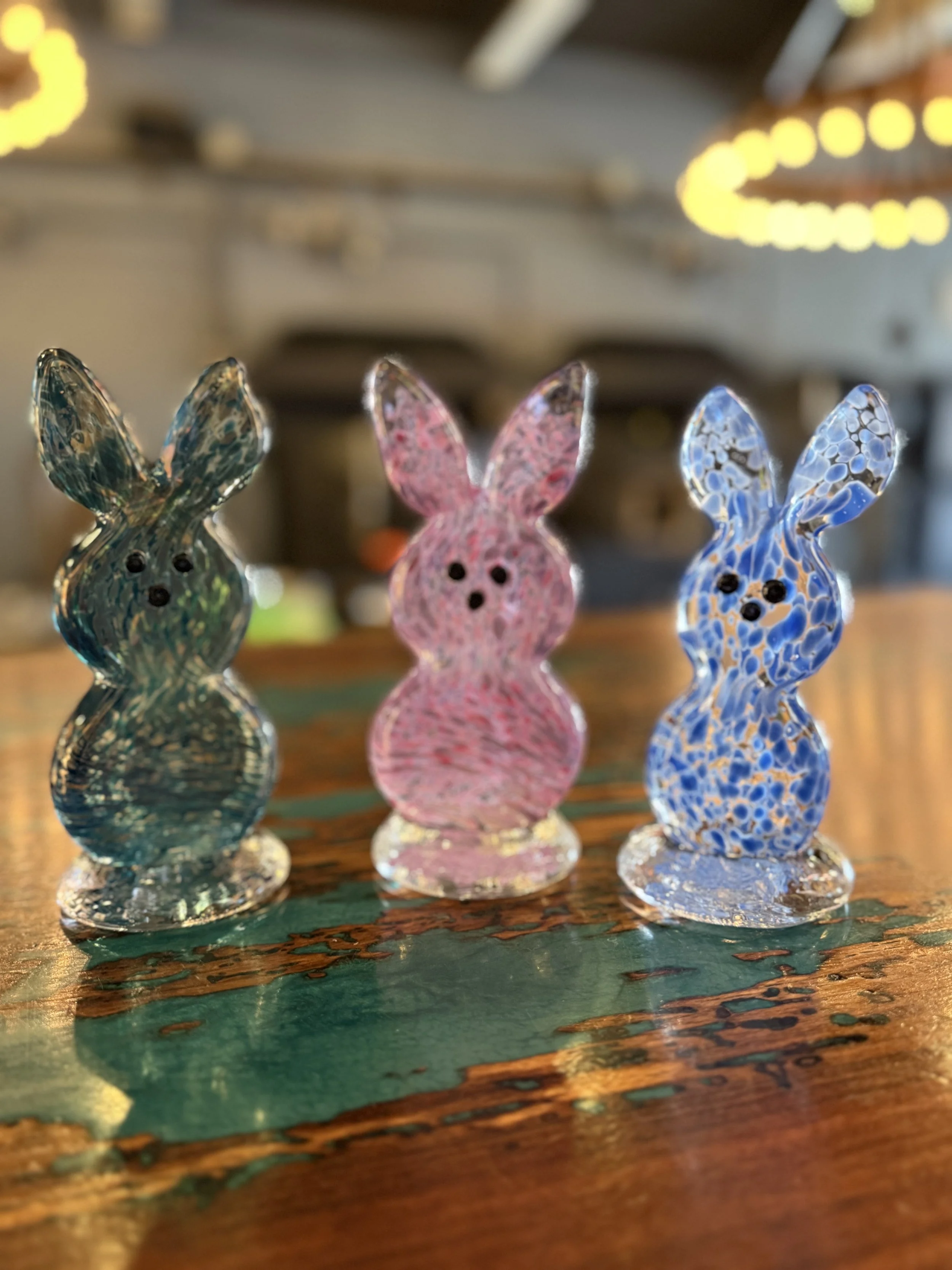 Three colorful glass bunny figurines with black eyes, placed on a wooden surface with a blurred background.