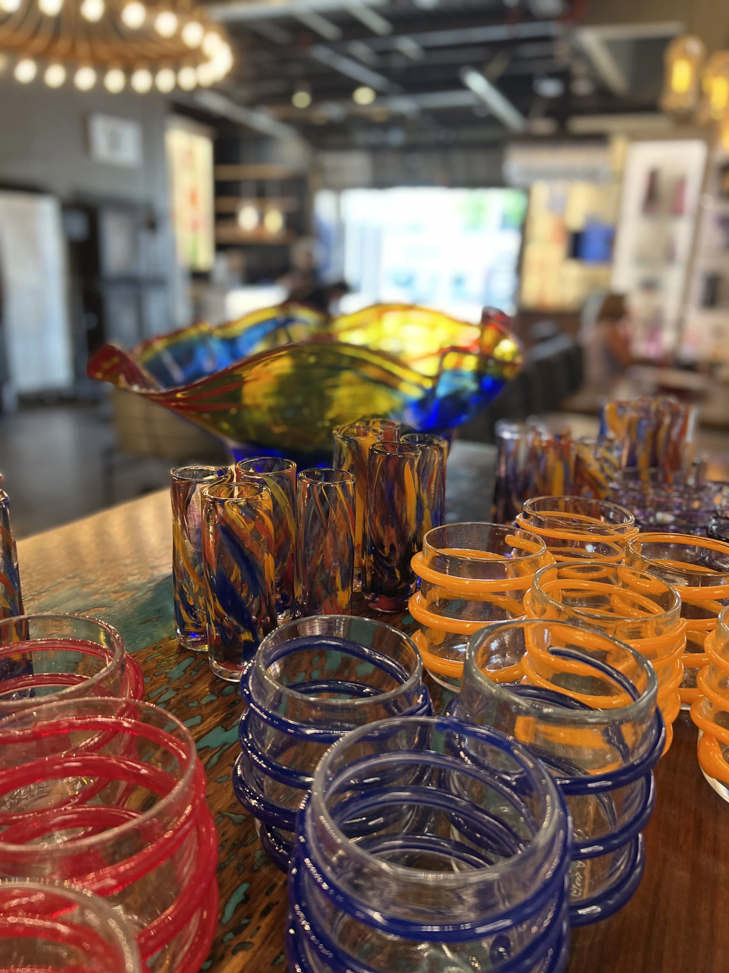 Colorful glass bowls and tumblers at glass studio, displayed on a wooden table with a blurred background of store interior.