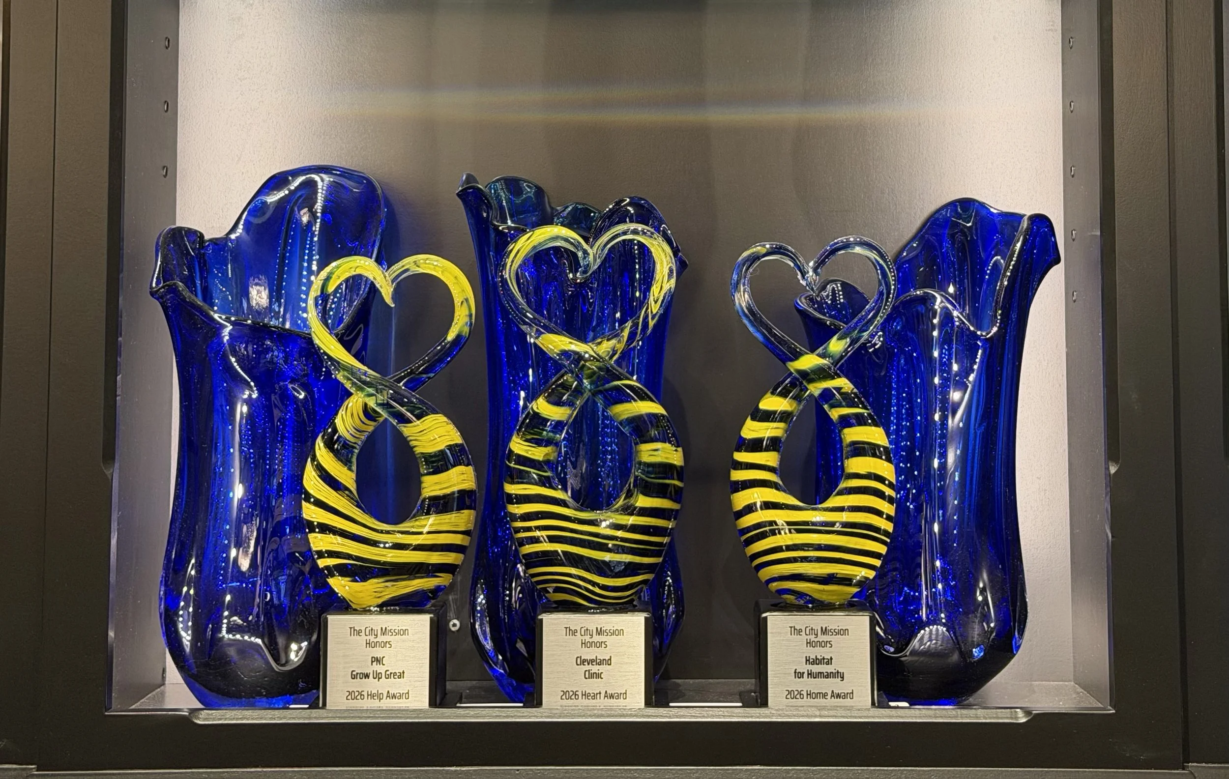 Three blue glass art awards with yellow and black striped heart-shaped accents, displayed in a glass case. Each award has a plaque with different honors and descriptions.
