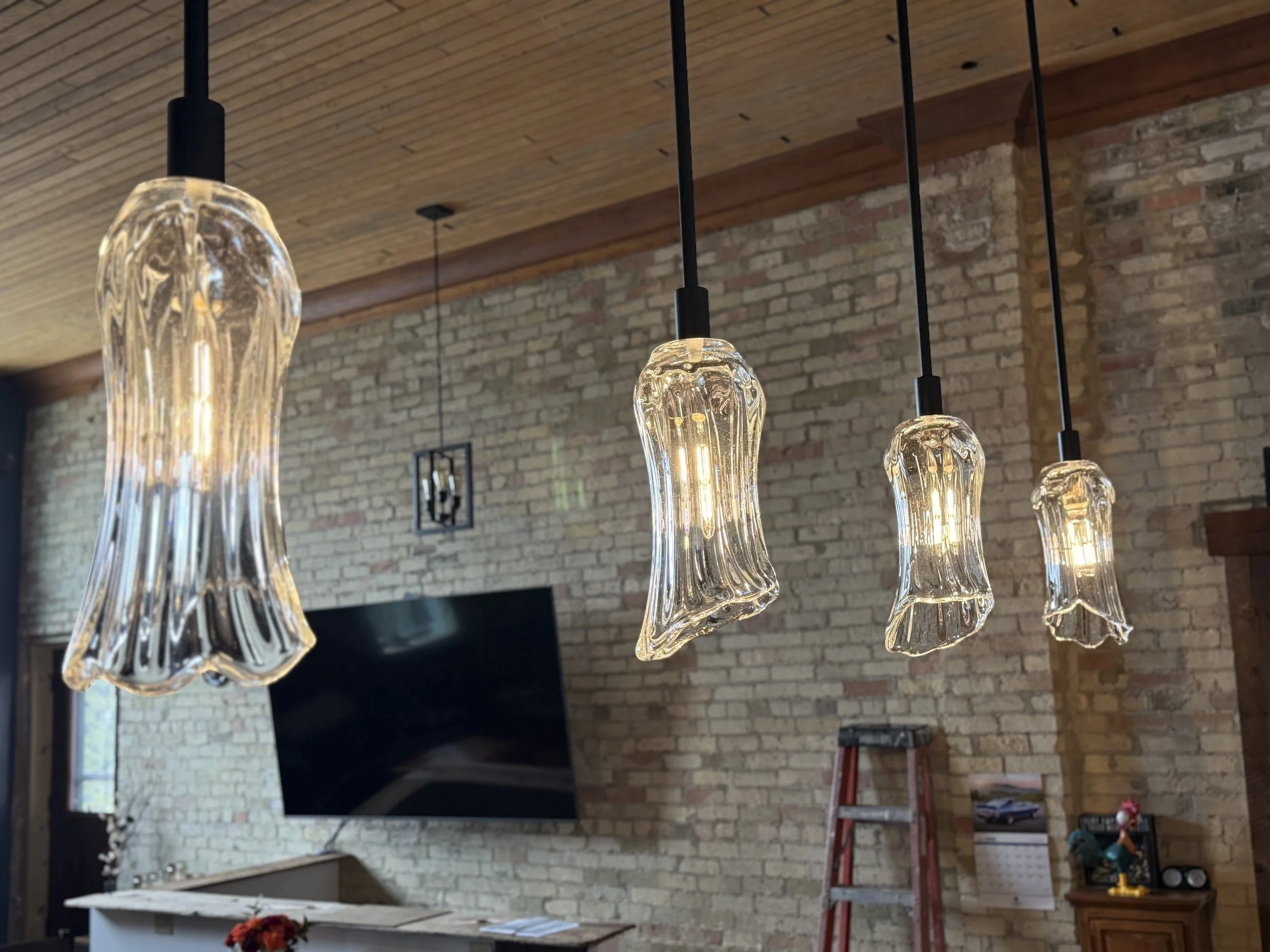 Four hanging glass pendant lights with decorative, wavy shapes in a cozy living room with an exposed brick wall and a wooden ceiling.