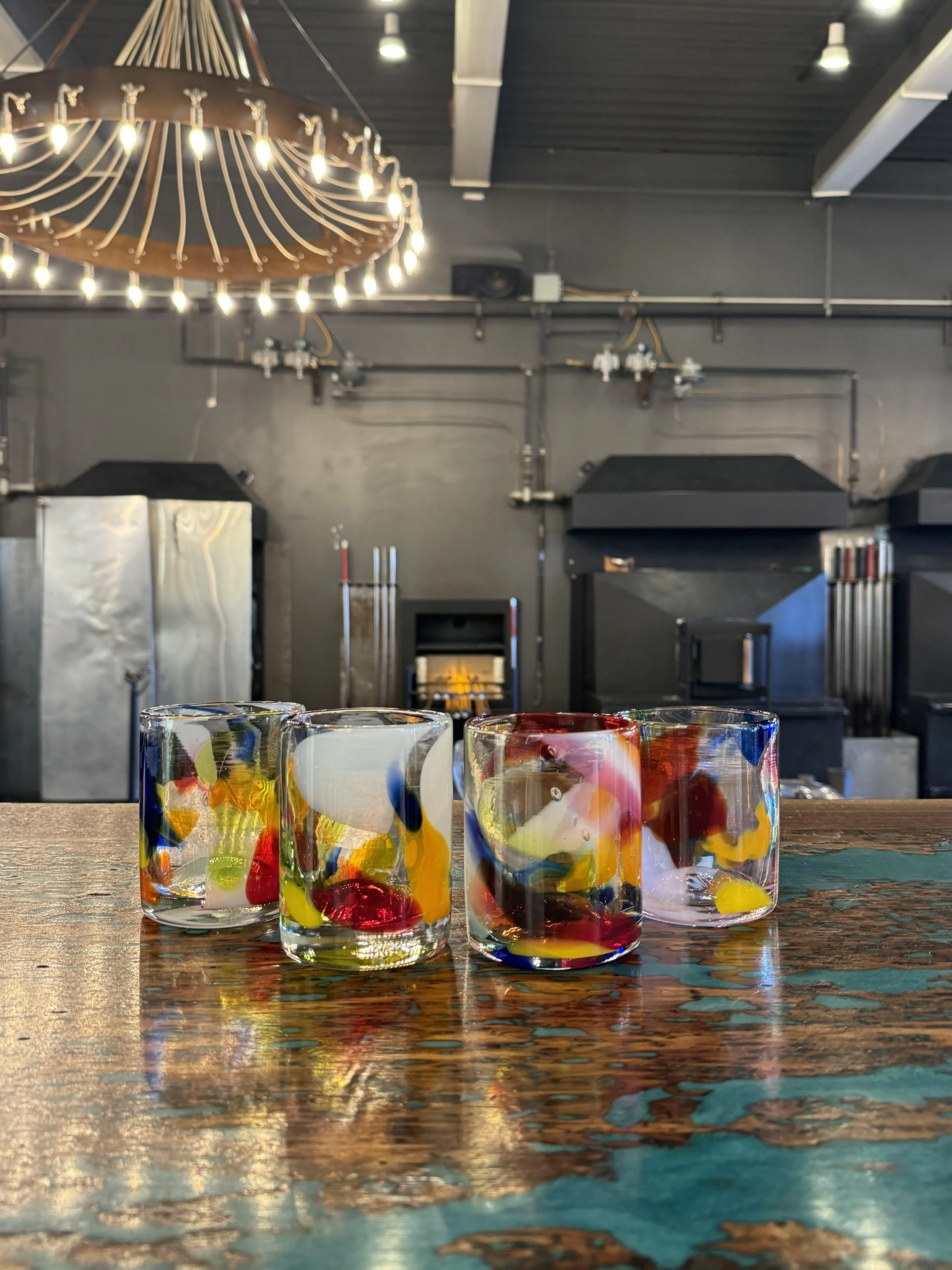 Four colorful glass tumblers with abstract designs on a wooden table inside a modern industrial-style space, with a large chandelier hanging from the ceiling in the background.