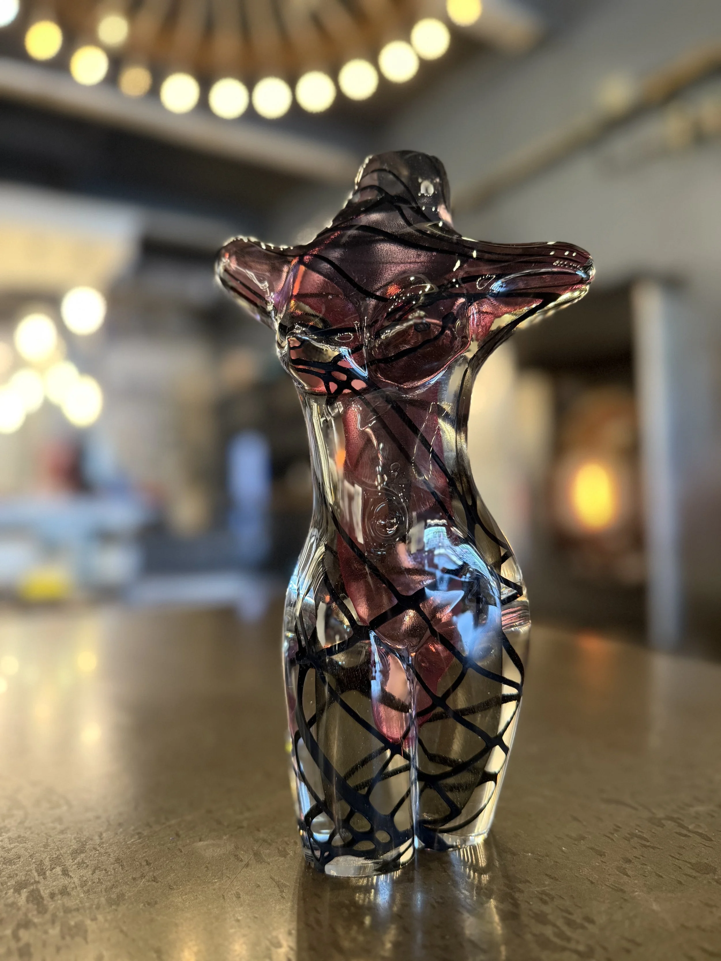 Colorful glass sculpture resembling a female torso figure, decorated with black swirling lines, set on a tabletop in a warmly lit indoor studio.