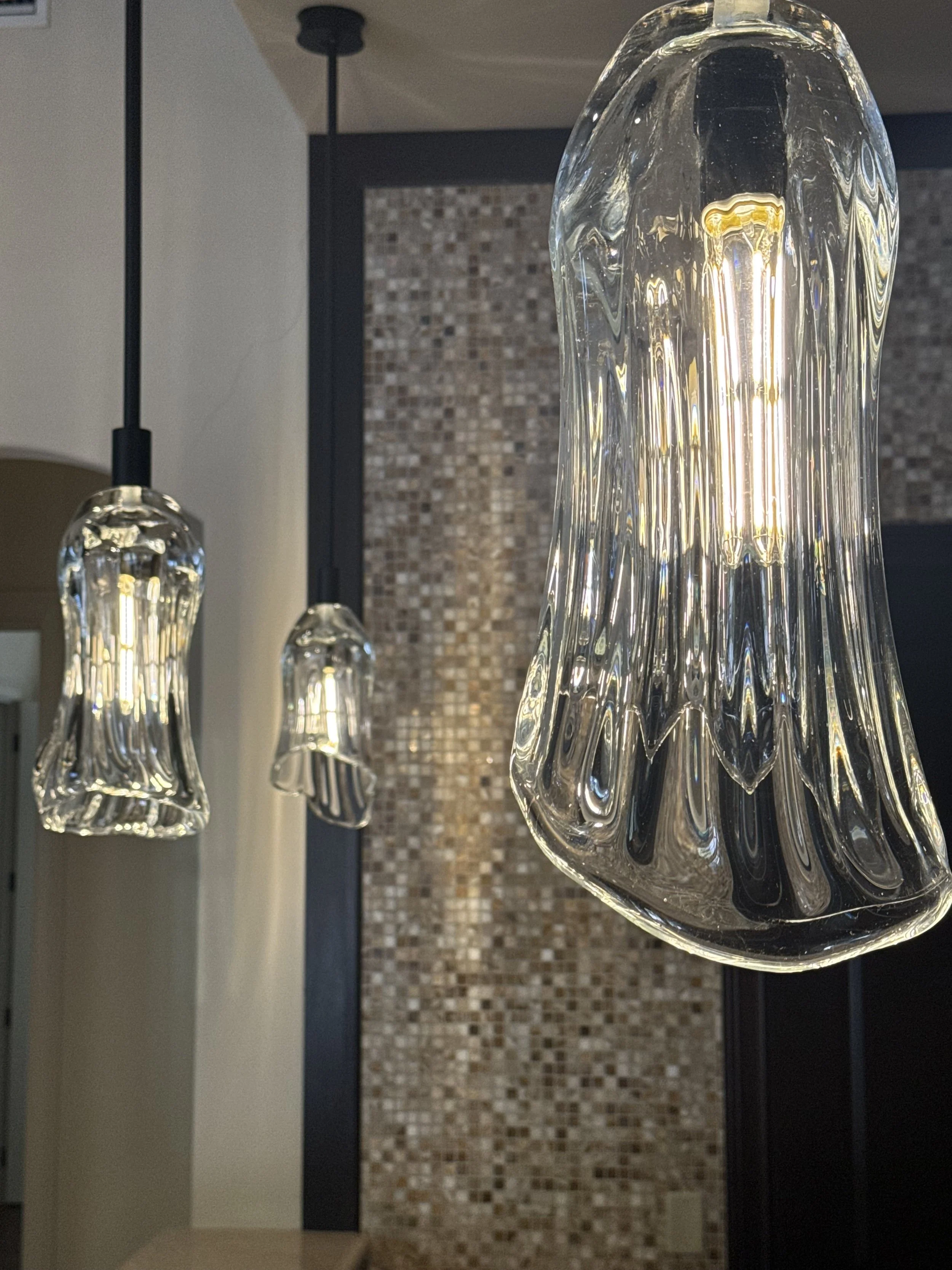Three hanging glass pendant lights, emitting warm light, in front of a textured mosaic tile wall inside a room.