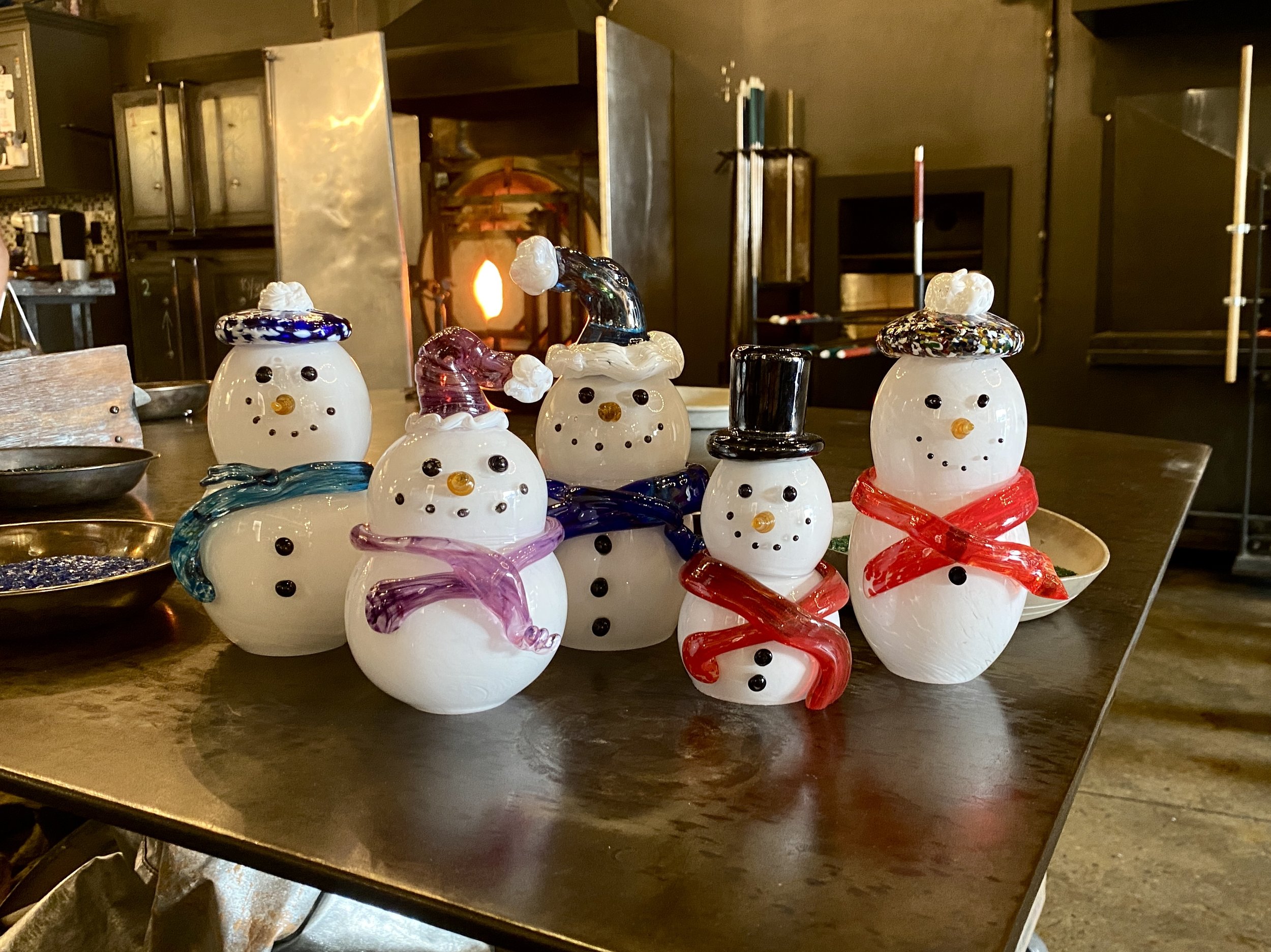Set of six glass snowman decorations with colorful scarves and hats arranged on a wooden table, with a glassblowing furnace in the background.