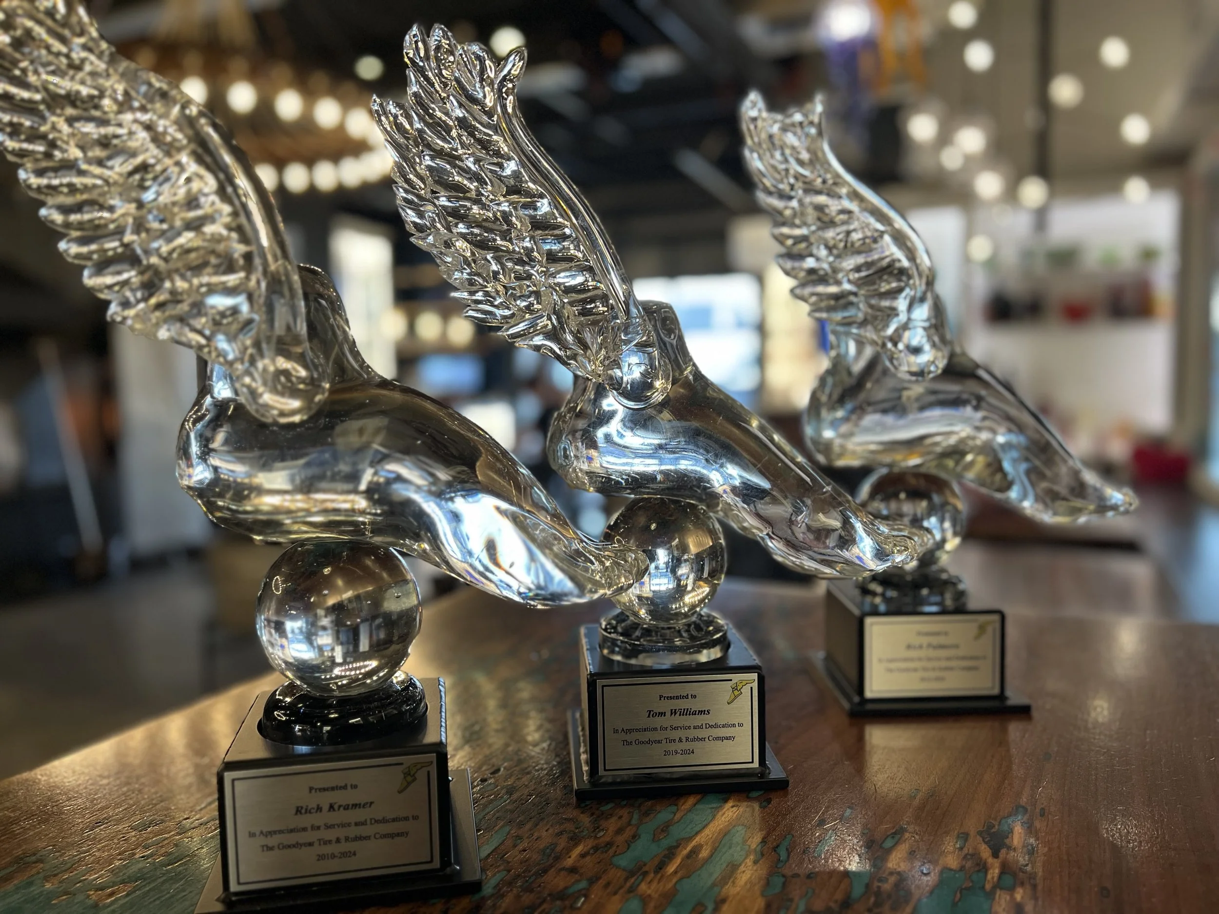 Three glass awards shaped like feet with wings, mounted on spherical bases and black plaques, displayed on a wooden table in a decorated glassblowing studio.