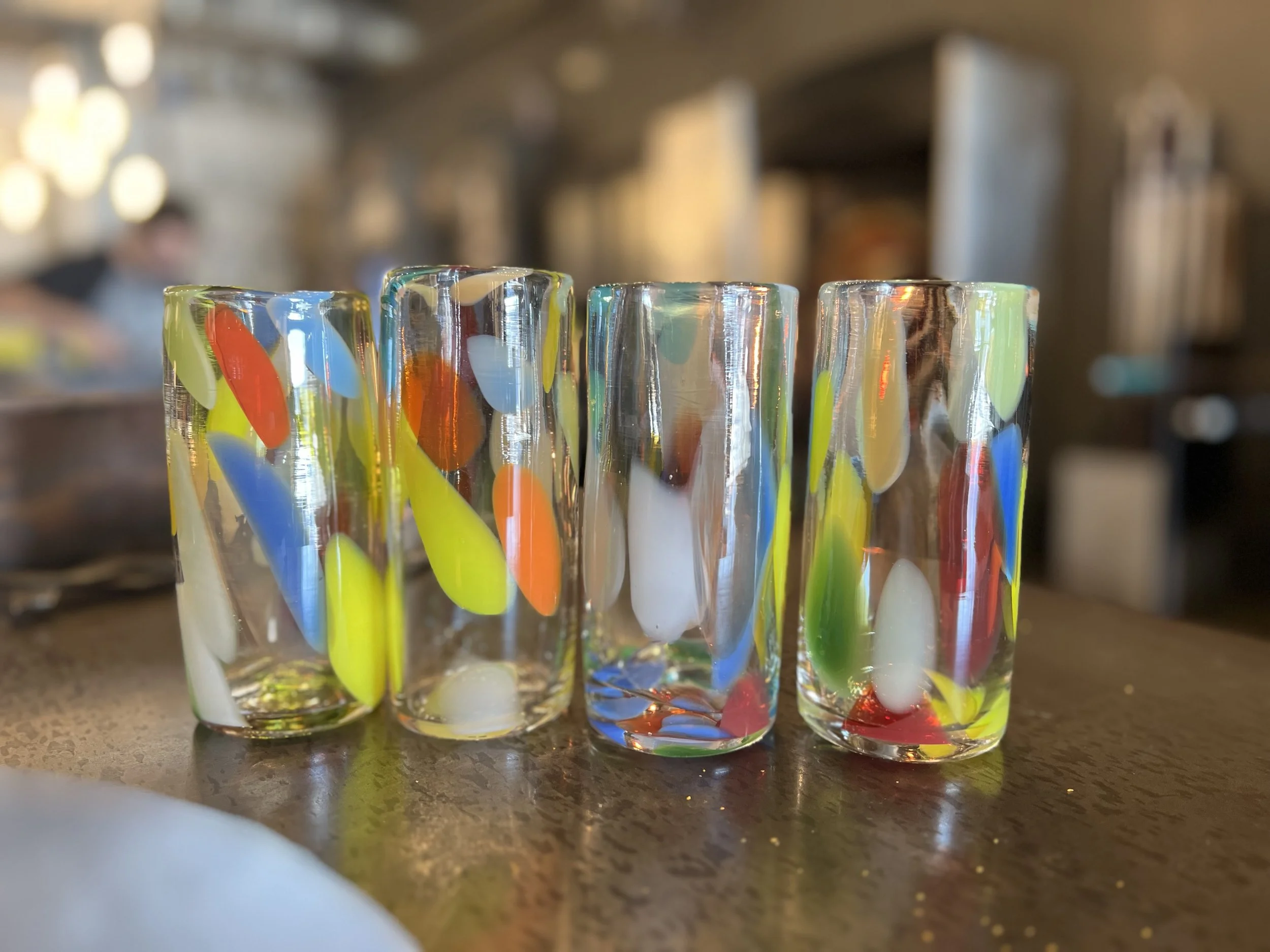 Four colorful glass tumblers with multicolored abstract patterns on a wooden surface, blurring into background with a person in an industrial setting