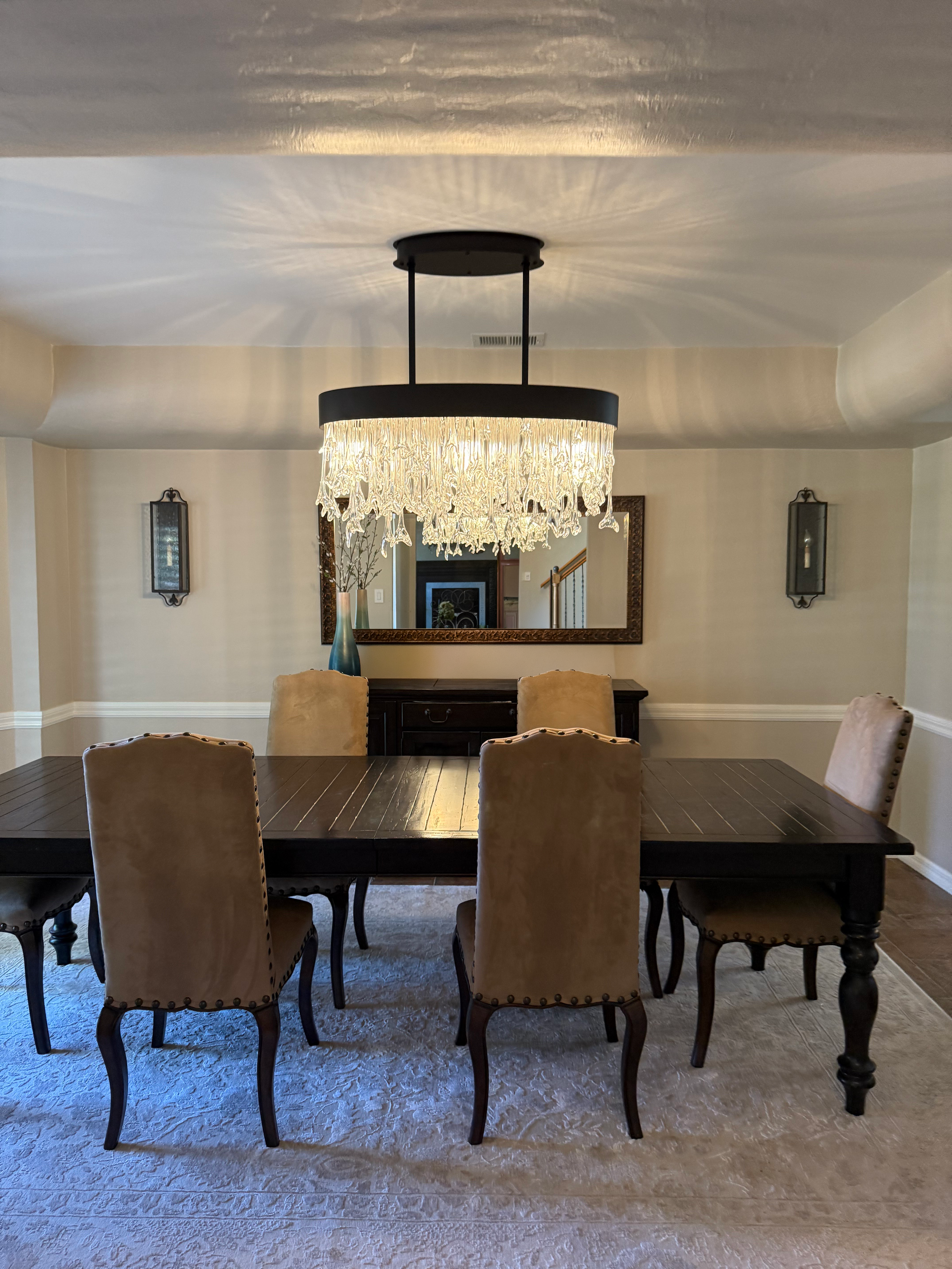Dining room with a dark wooden table, upholstered beige chairs, a mirror on the wall, a chandelier hanging from the ceiling, and decorative wall sconces.