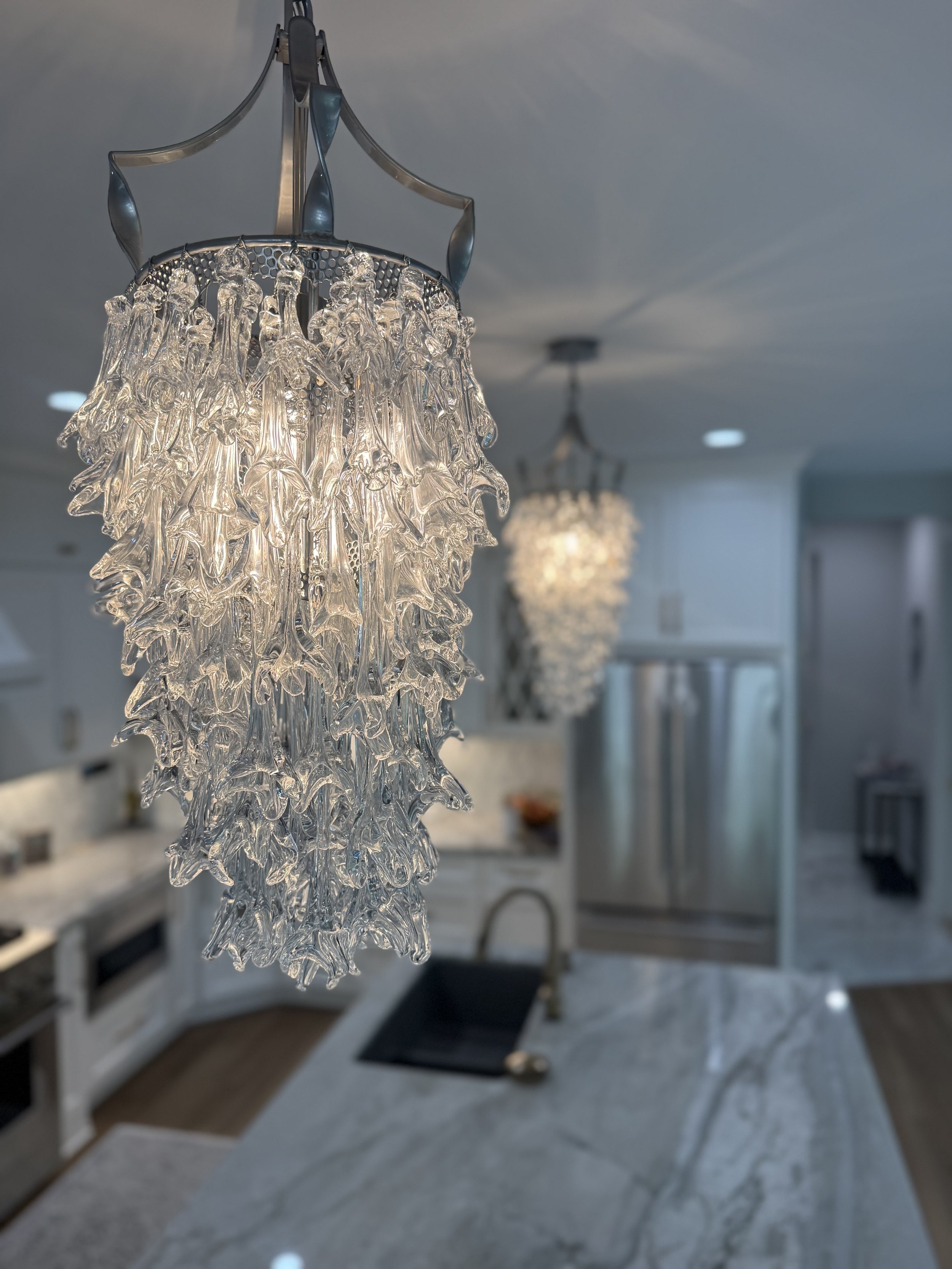 Close-up of a decorative glass chandelier hanging in a modern kitchen with white cabinetry and marble countertops.