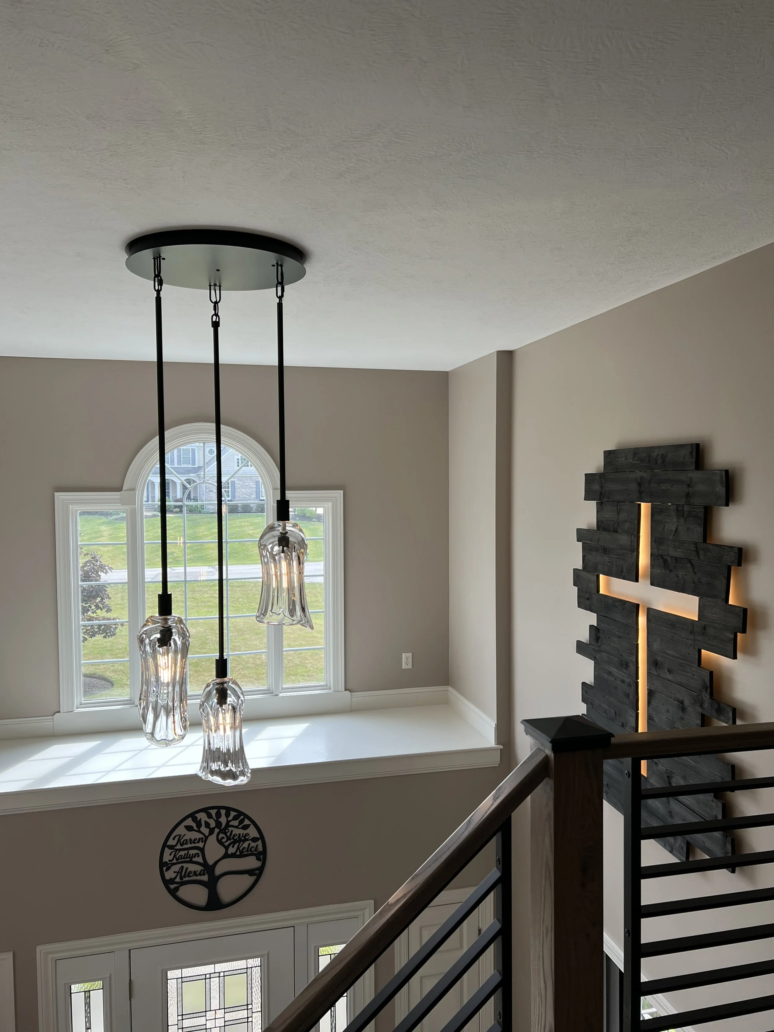 Interior view of a home staircase landing with a large window, sleek black and glass pendant light fixture, a black decorative cross wall art with backlighting.