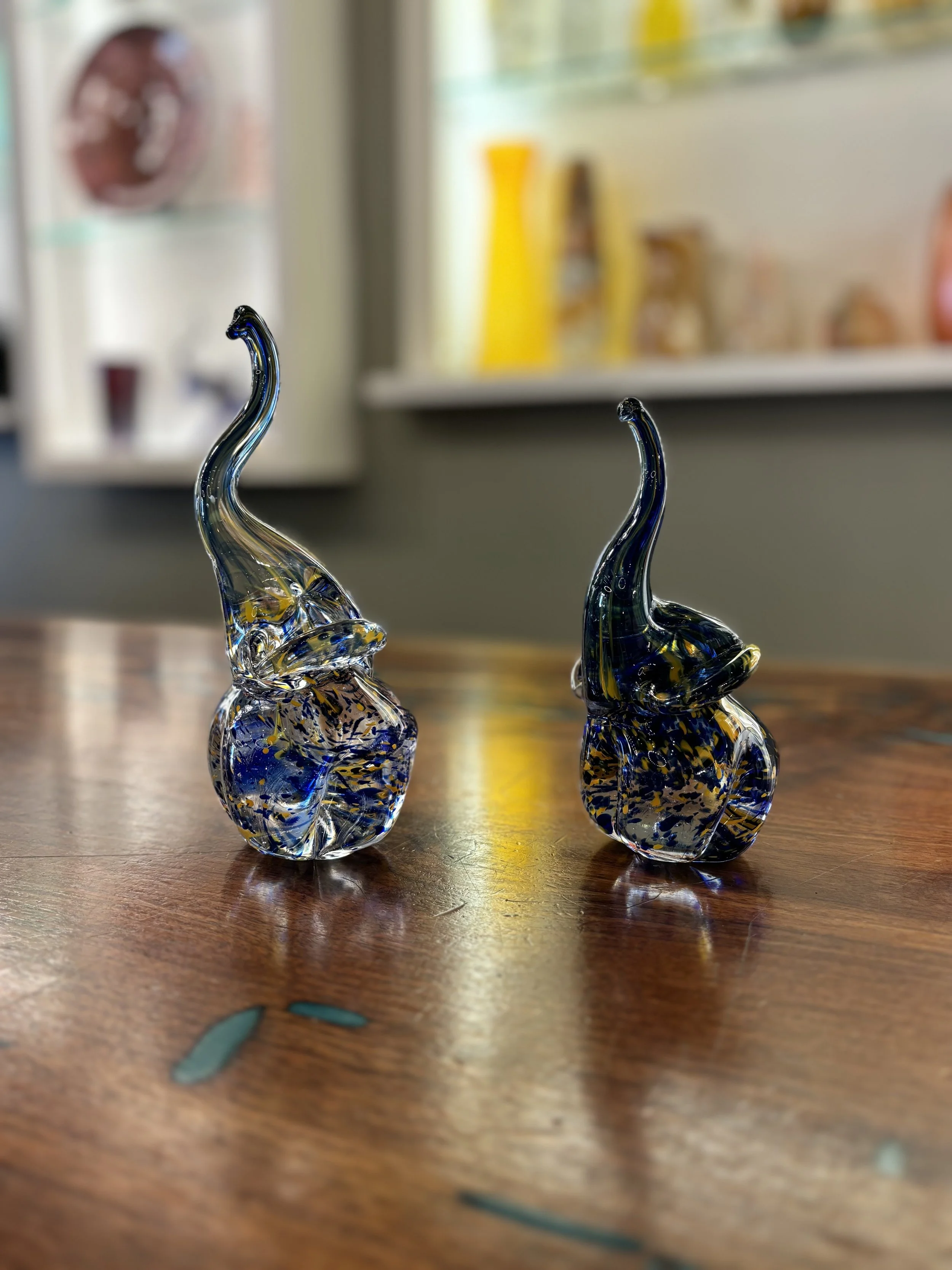 Two glass elephant sculptures with elongated trunks are displayed on a wooden surface in a room, with a background of blurred colorful objects on a shelf.