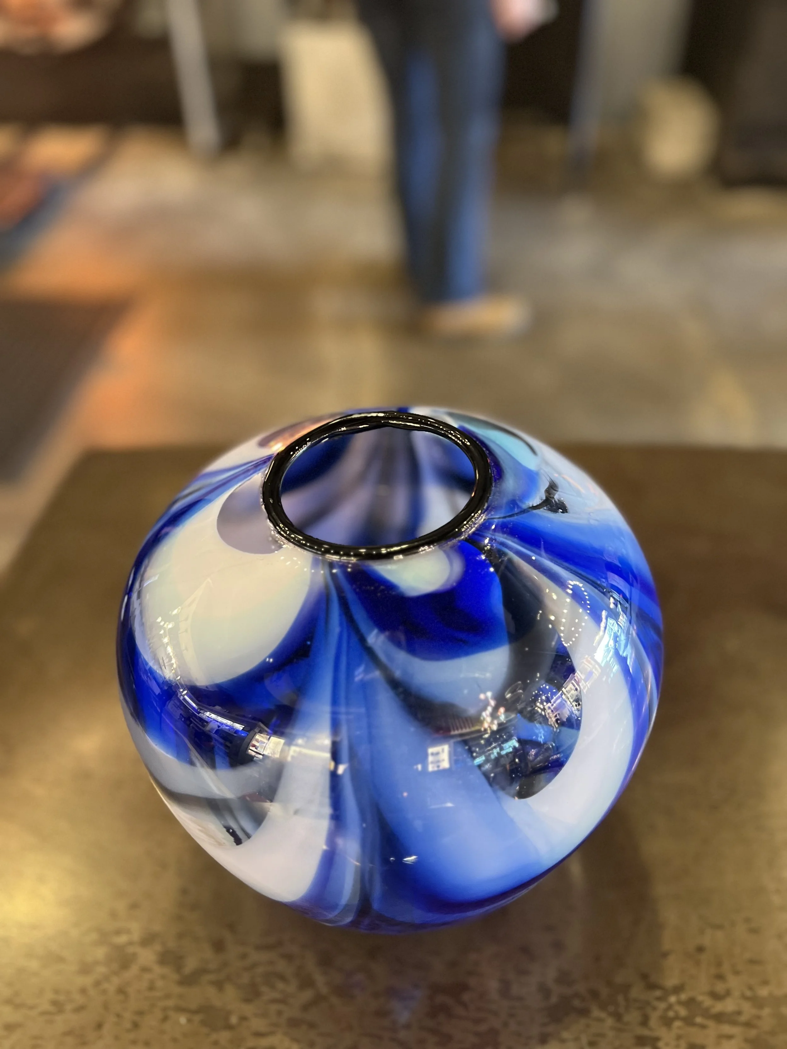 Close-up of a blue and white marble glass vase on a wooden surface, with a blurred person and interior background.