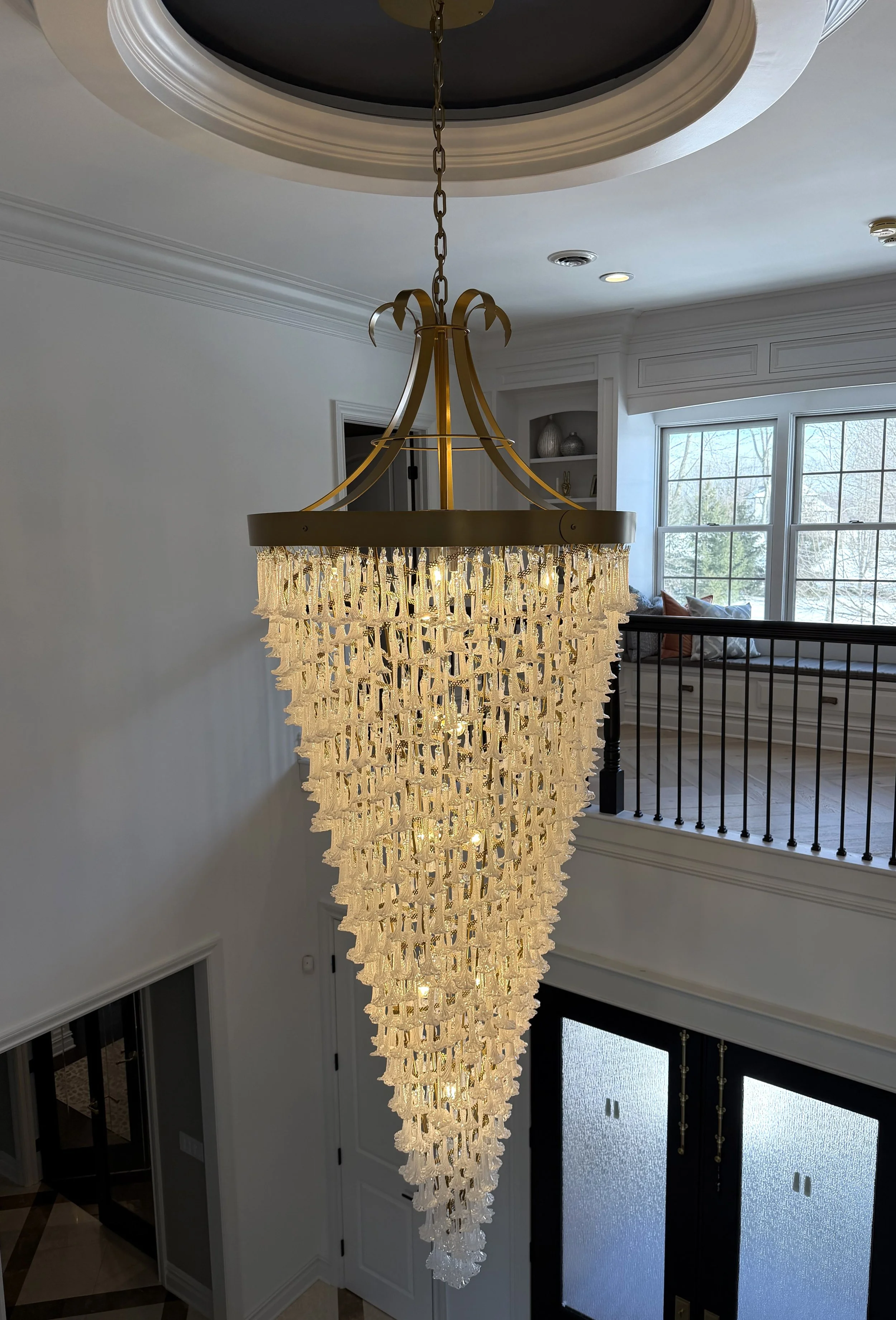 A large, elegant chandelier hanging from a ceiling in a spacious, well-lit home interior with a view of a living area and large window.