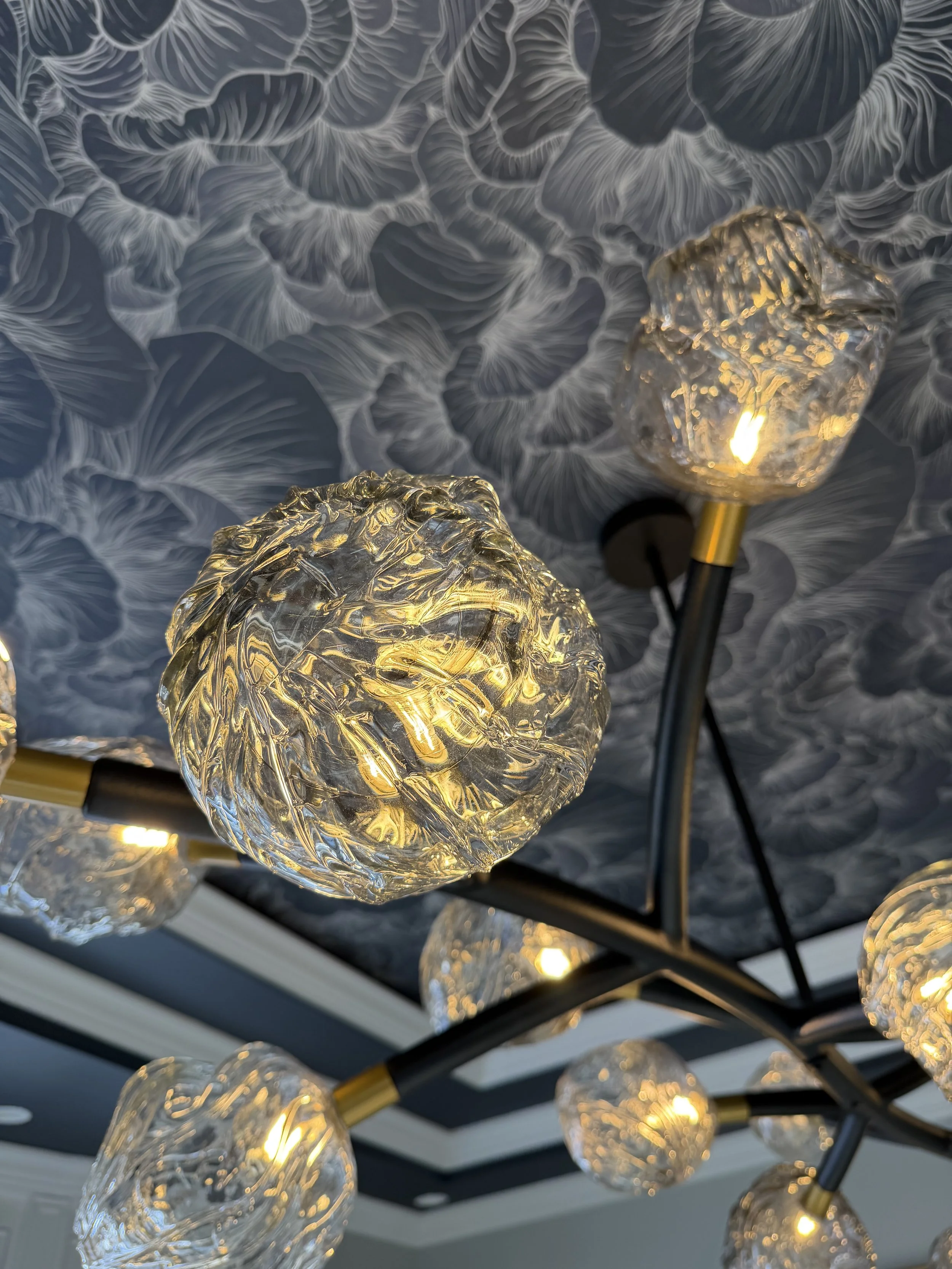 Close-up of a modern chandelier with textured glass globes and warm yellow lights, hanging from a ceiling with dark floral-patterned wallpaper.